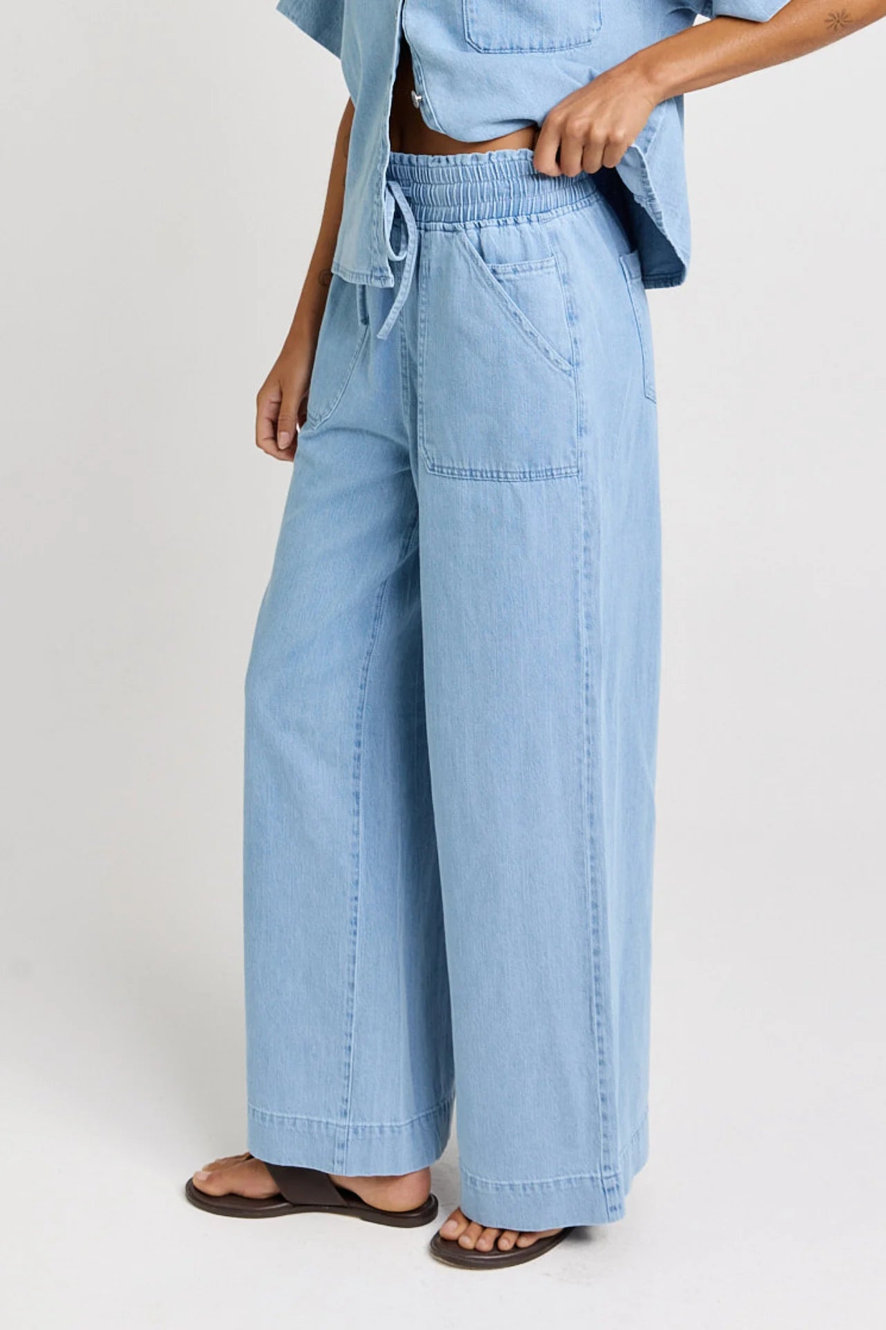 Image of A woman wearing Betty Basics light blue wide-leg denim pants with an elastic drawstring waistband, paired with a matching light blue top and dark brown sandals, shown against a plain light gray studio background.