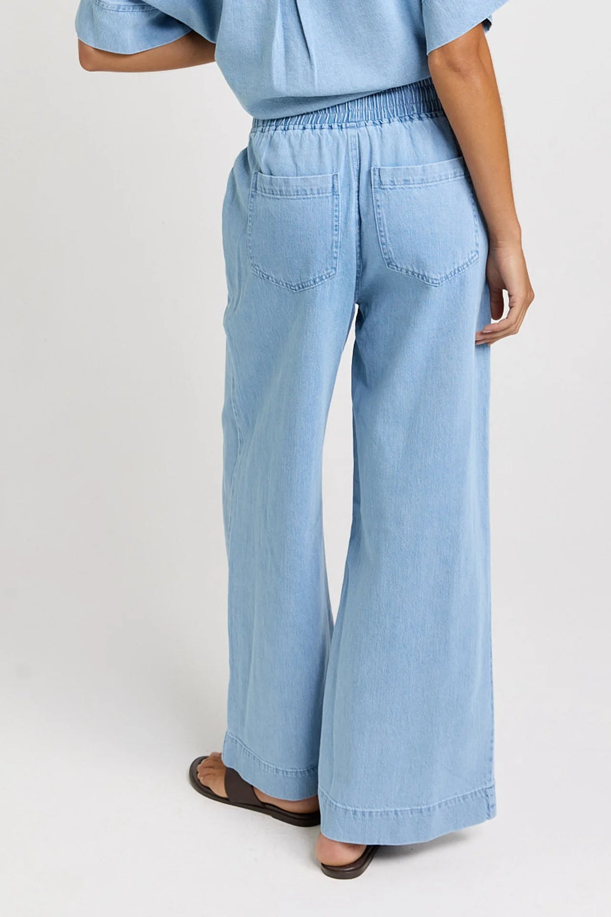 A woman shown from the back wearing Betty Basics light blue wide-leg denim pants with an elastic waistband and back pockets, paired with a matching light blue top and brown sandals, against a plain light gray studio background.