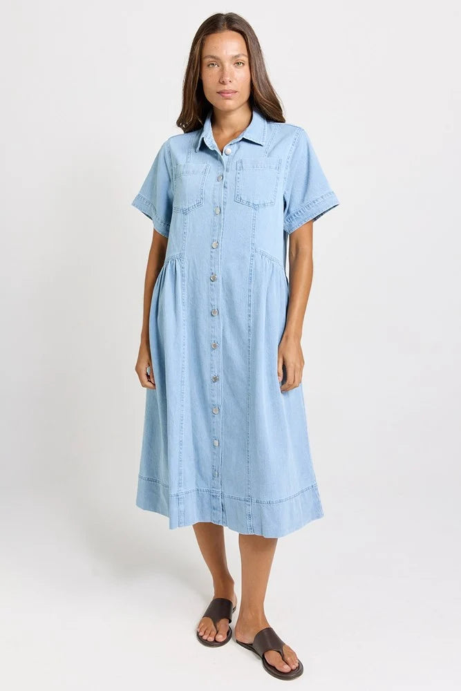 Image of A woman modeling a Betty Basics light blue denim shirt dress with short sleeves, a button-down front, and a relaxed fit, paired with dark brown slide sandals against a plain white studio background.