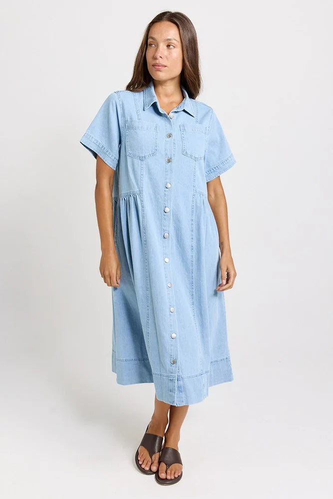 Image of A woman modeling a Betty Basics light blue denim short-sleeve button-down midi dress with a relaxed, tiered silhouette, paired with brown leather slide sandals, against a plain light gray studio background.