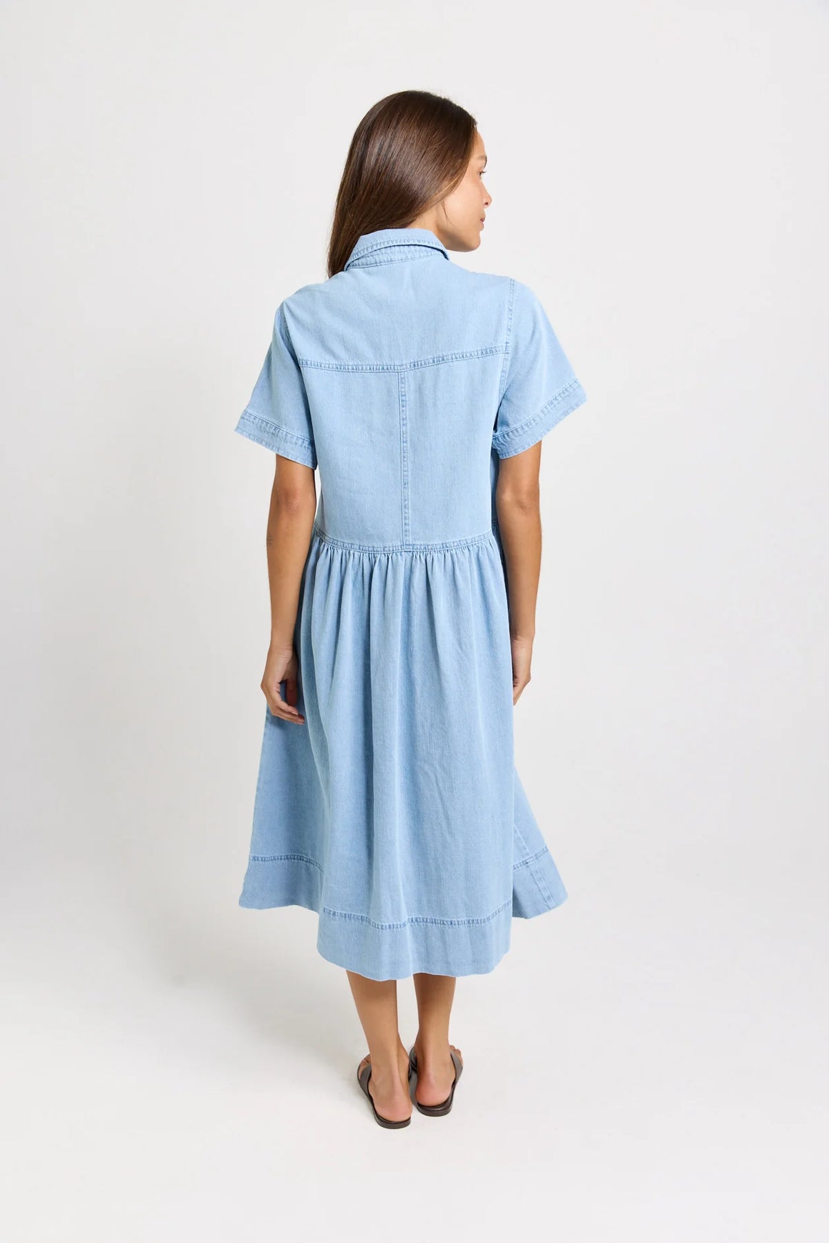 A woman viewed from the back wearing a Betty Basics light blue short-sleeve midi dress with a gathered waist, shown against a plain light gray studio background.