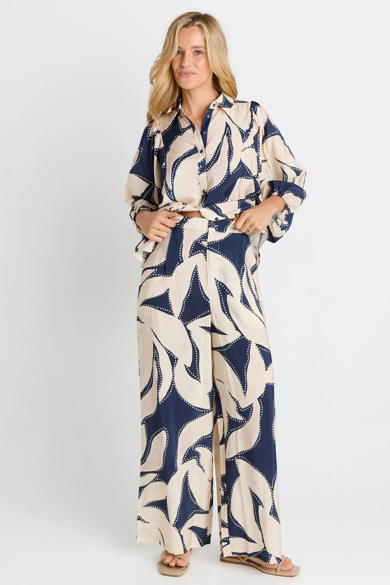 Image of A woman modeling a Betty Basics long-sleeve, wide-leg jumpsuit with a navy blue and cream abstract leaf pattern, standing against a light gray studio background.