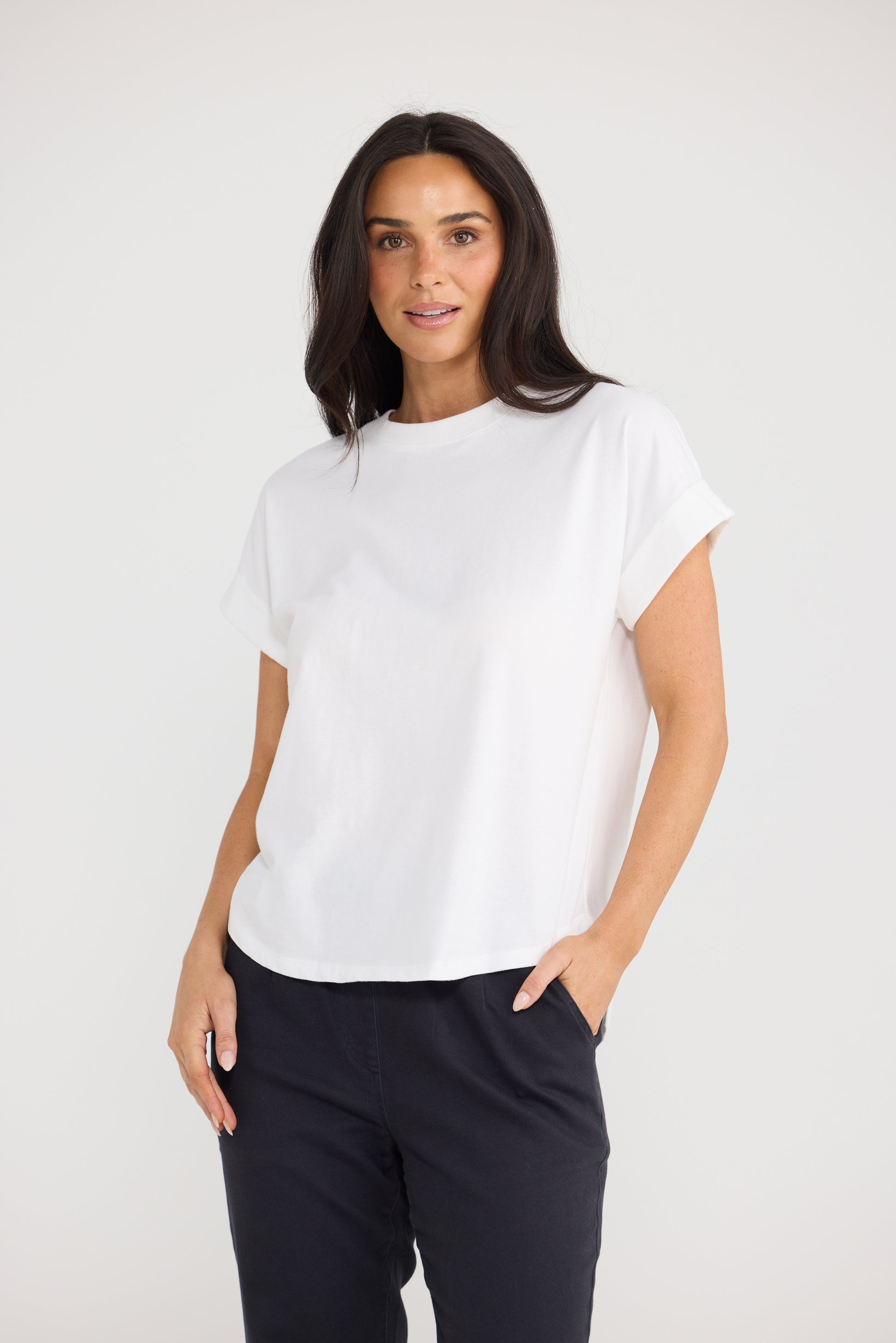 A woman modeling a Brave & True white short-sleeve crewneck t-shirt paired with black pants, standing against a plain light gray studio background.