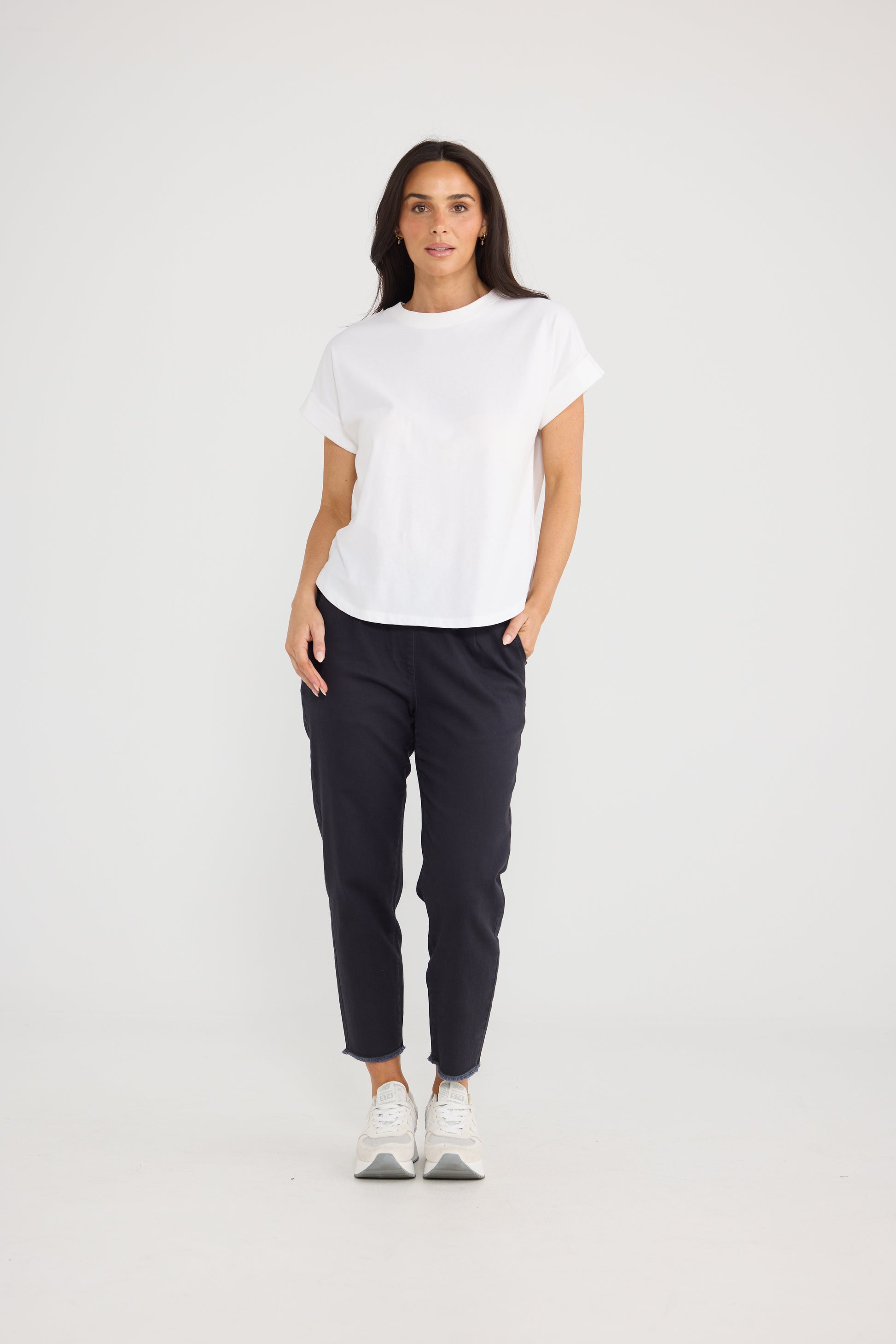 A woman modeling a Brave & True white short-sleeve crewneck t-shirt and dark navy cropped pants with white sneakers, standing against a seamless white studio background.