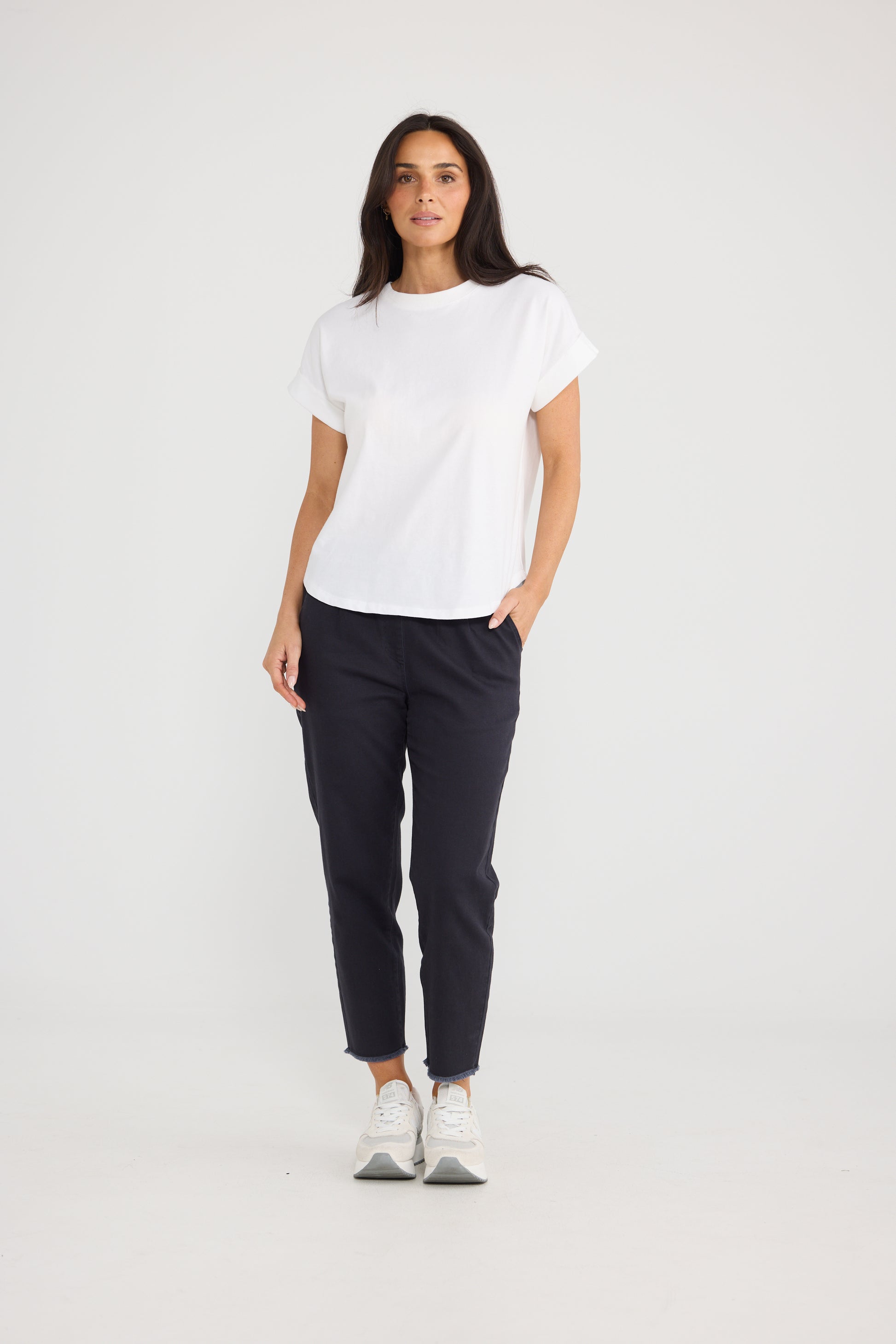 A woman modeling a Brave & True white short-sleeve crewneck t-shirt and dark navy cropped trousers, paired with white sneakers, against a plain light gray studio background.
