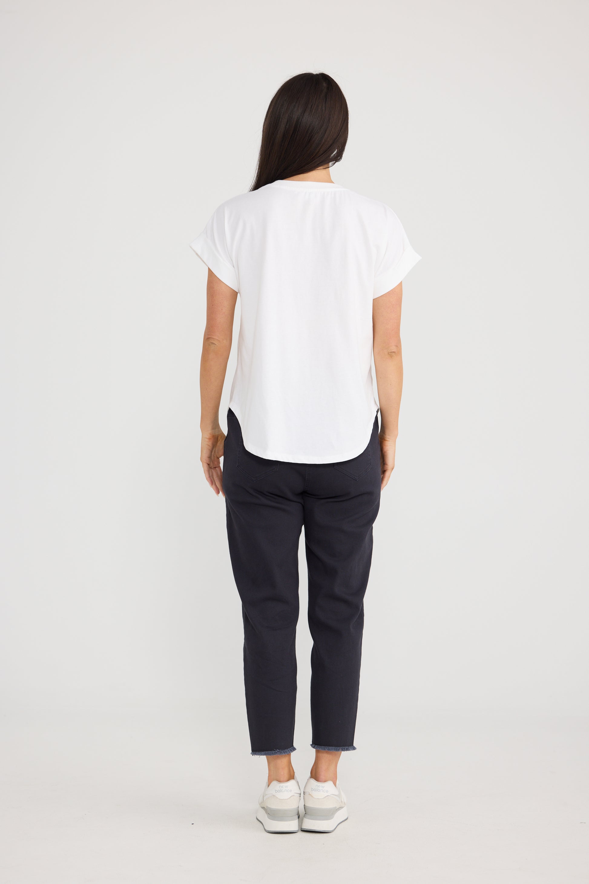 A woman viewed from behind wearing a Brave & True white short-sleeve top, dark navy pants, and white sneakers against a plain light gray studio background.