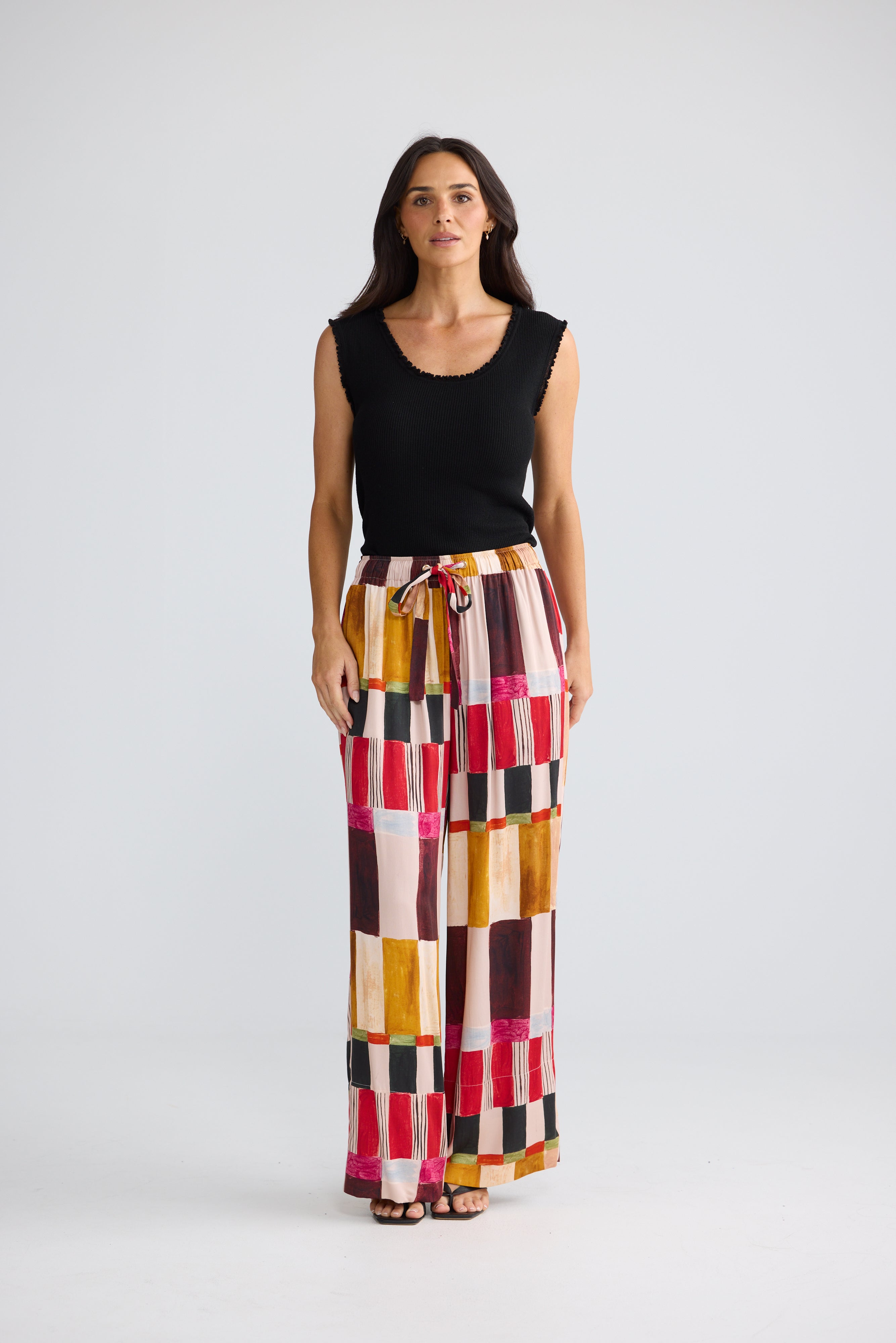 Image of A woman modeling a sleeveless black top and high-waisted, Brave & True wide-leg trousers featuring a colorful geometric patchwork pattern of pink, yellow, orange, brown, and black blocks, set against a plain light gray studio background.