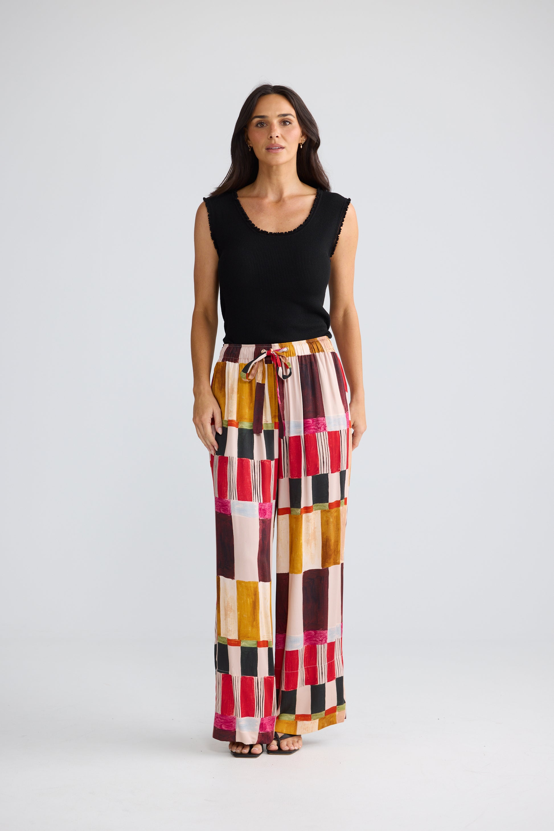 A woman modeling a sleeveless black top and high-waisted, Brave & True wide-leg trousers featuring a colorful geometric patchwork pattern of pink, yellow, orange, brown, and black blocks, set against a plain light gray studio background.