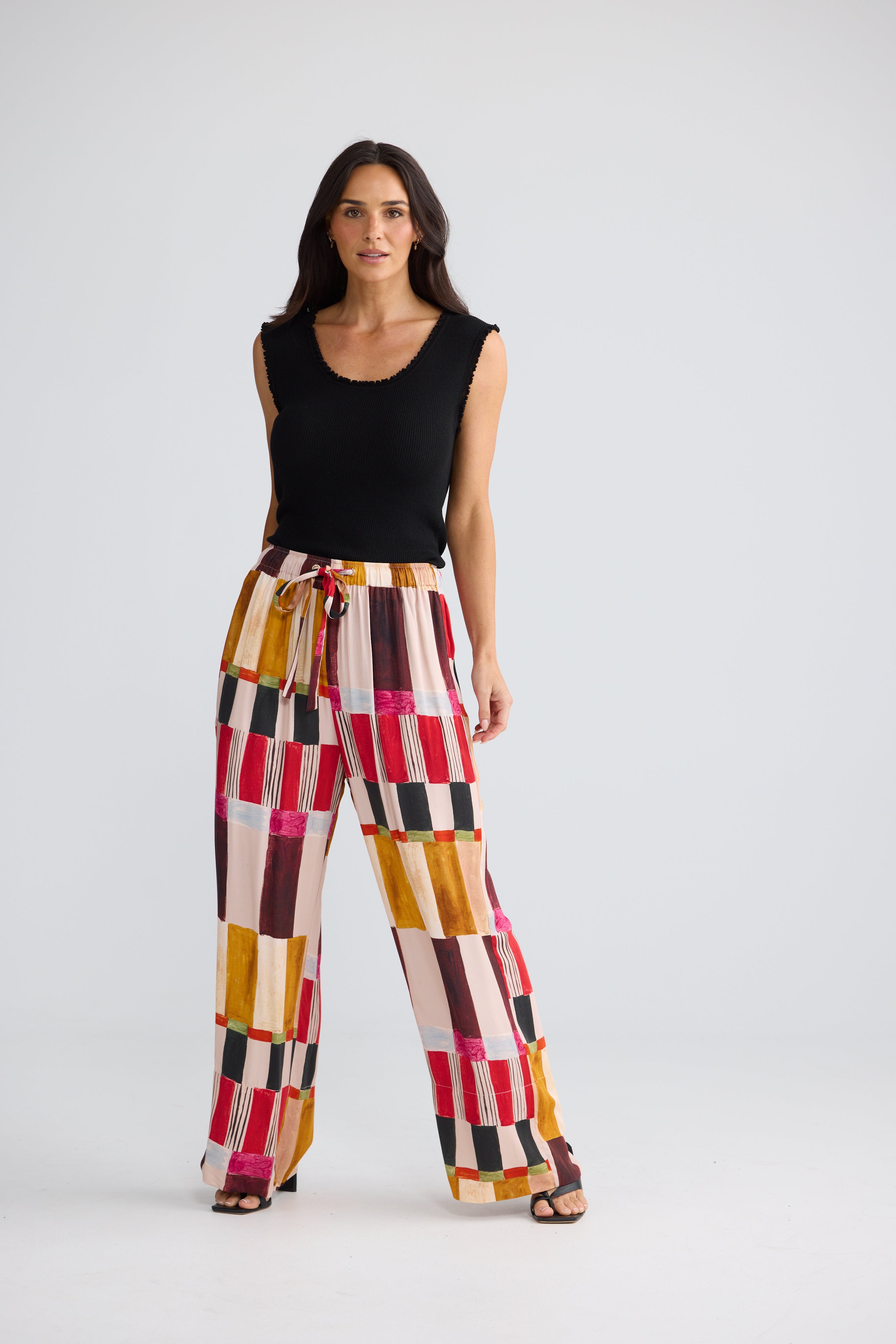 Image of A woman modeling a pair of Brave & True wide-leg pants with a colorful geometric patchwork pattern in pink, red, yellow, brown, and white, paired with a black sleeveless top, against a light gray studio background.