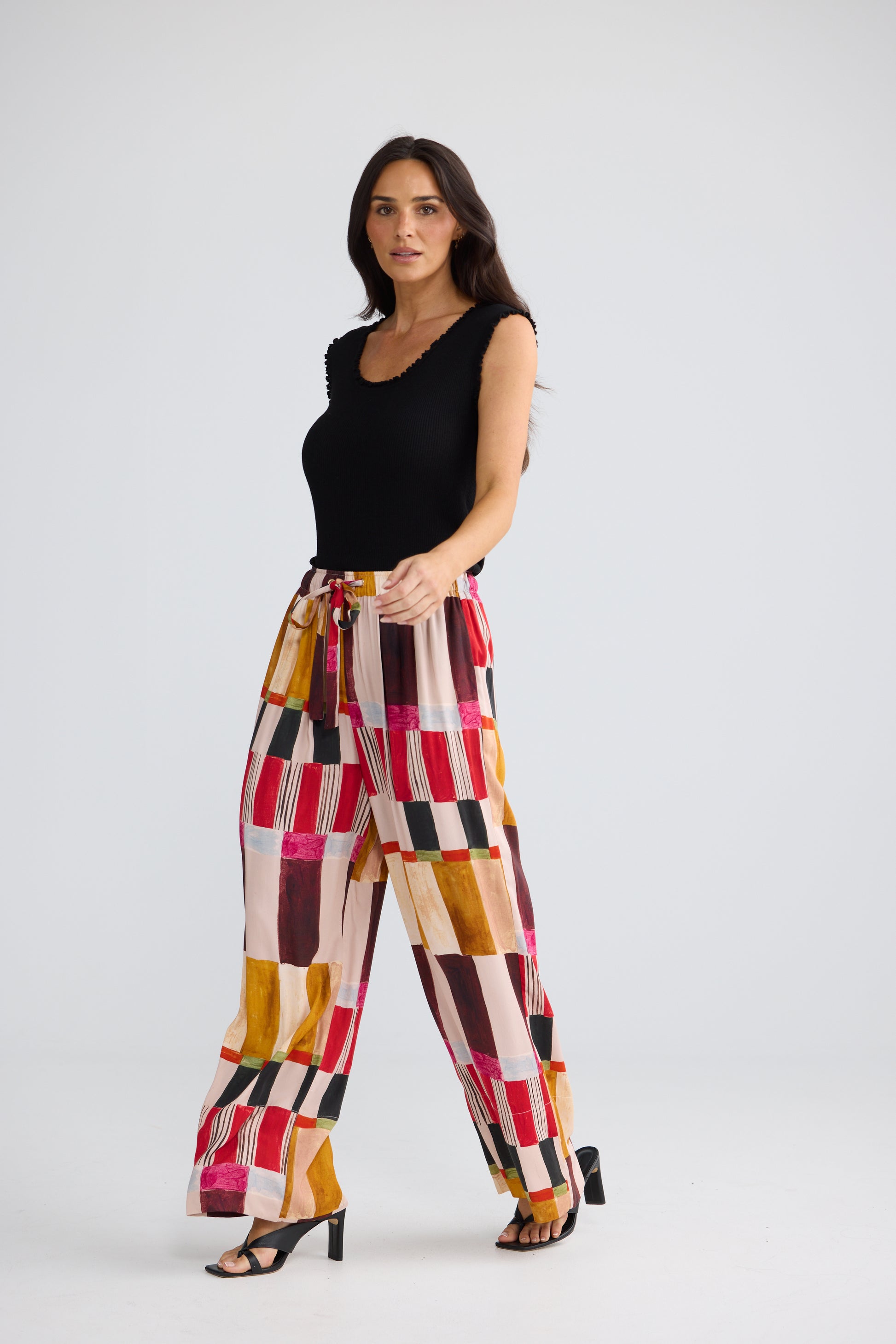 A woman modeling Brave & True high-waisted, wide-leg trousers with a colorful geometric block pattern in pink, orange, yellow, brown, and black, paired with a black sleeveless top and black heeled sandals, against a light gray studio background.