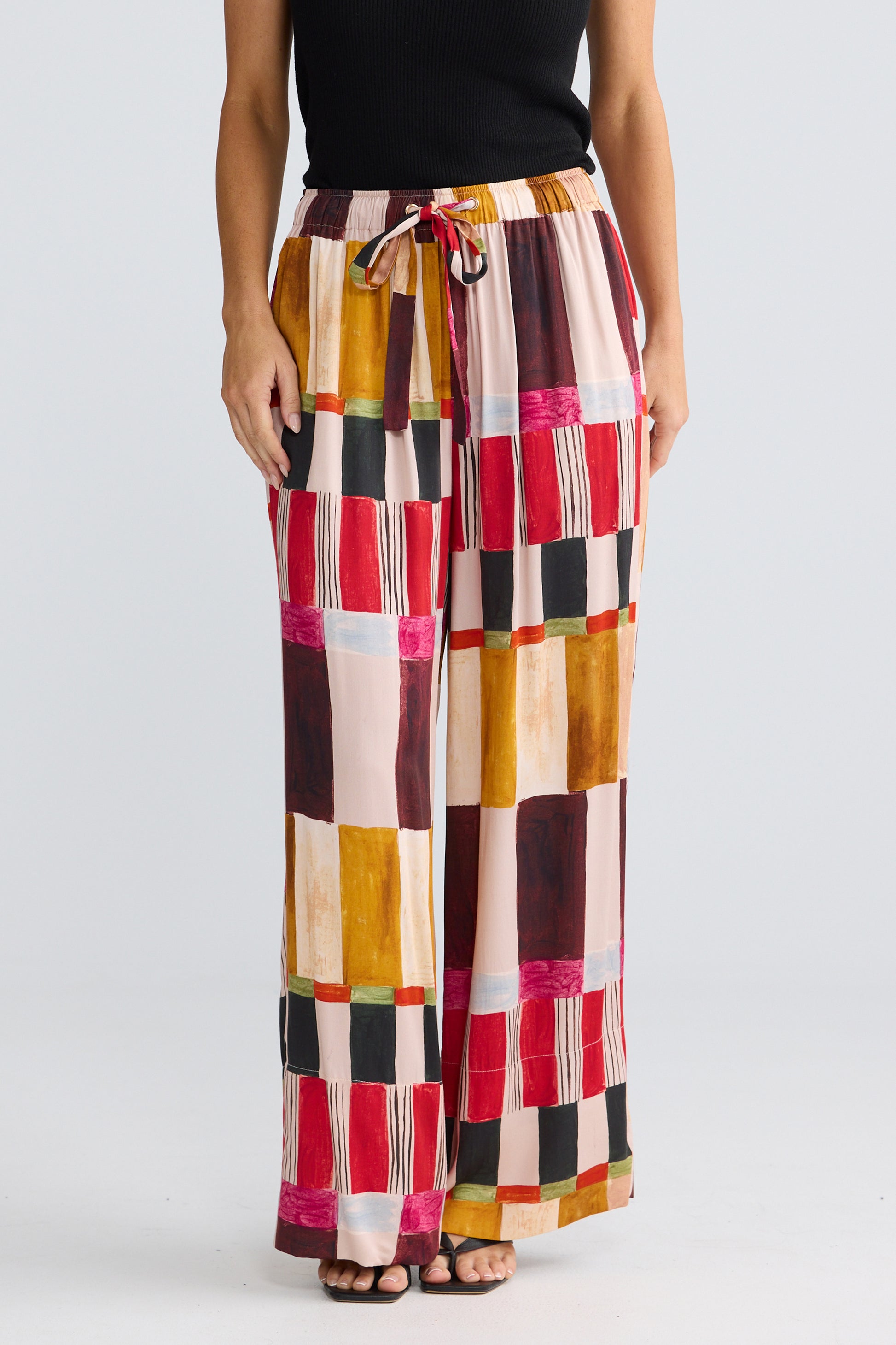 A model is shown from the waist down wearing Brave & True wide-leg trousers with a colorful patchwork print of red, yellow, brown, black, and pink blocks, paired with a black top and sandals.