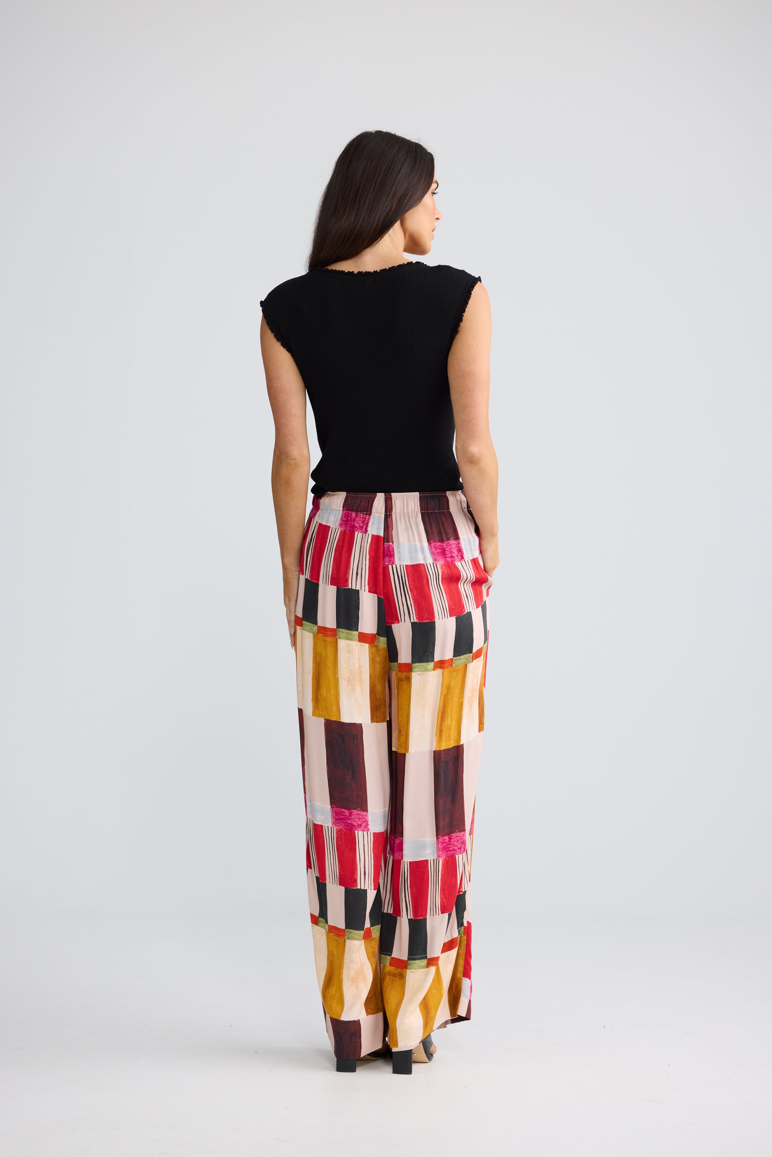 Image of A woman is shown from behind wearing a black sleeveless top and Brave & True high-waisted, wide-leg trousers with a colorful geometric block pattern in pink, yellow, brown, and white. She is photographed against a seamless light gray studio background.