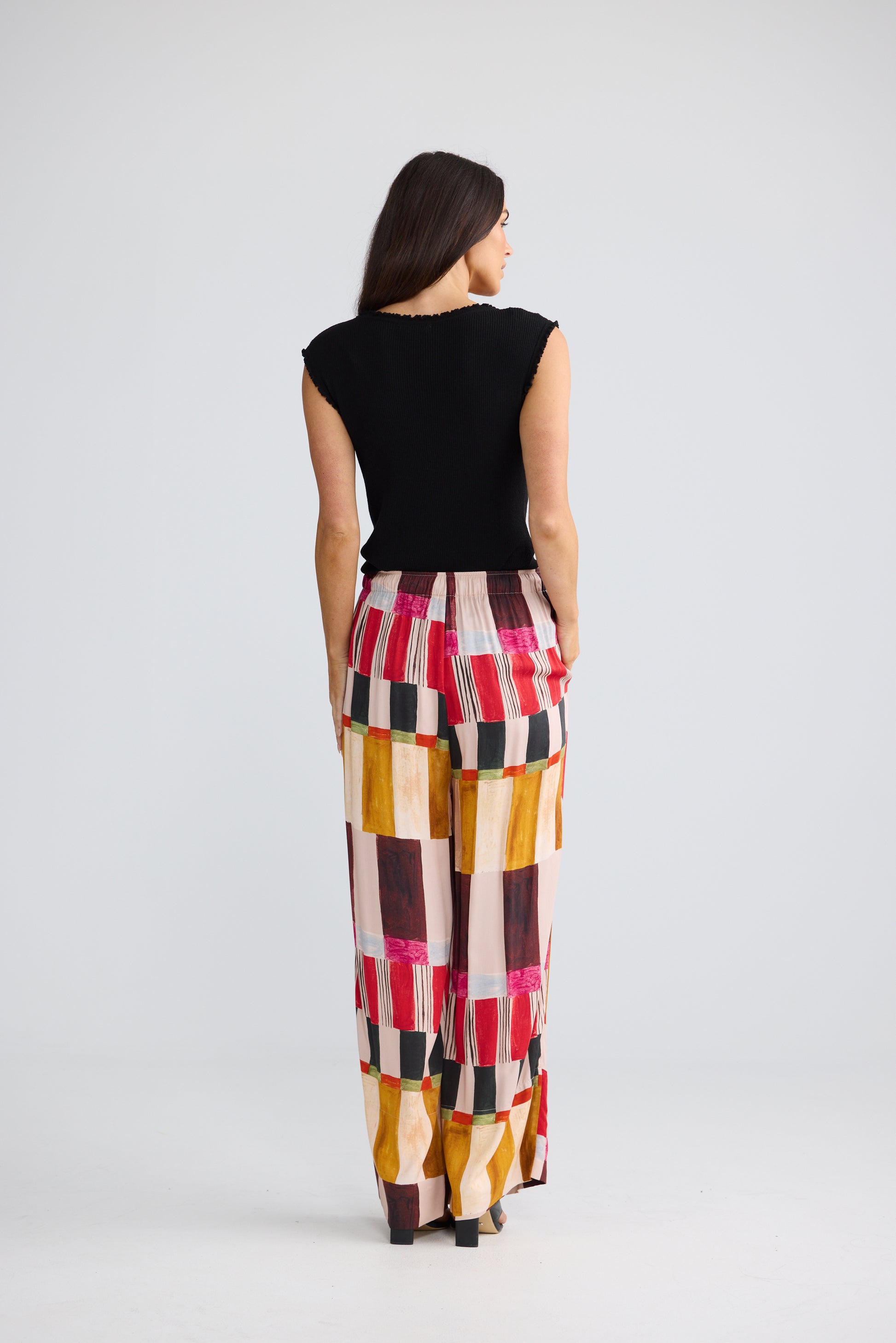 A woman is shown from behind wearing a black sleeveless top and Brave & True high-waisted, wide-leg trousers with a colorful geometric block pattern in pink, yellow, brown, and white. She is photographed against a seamless light gray studio background.