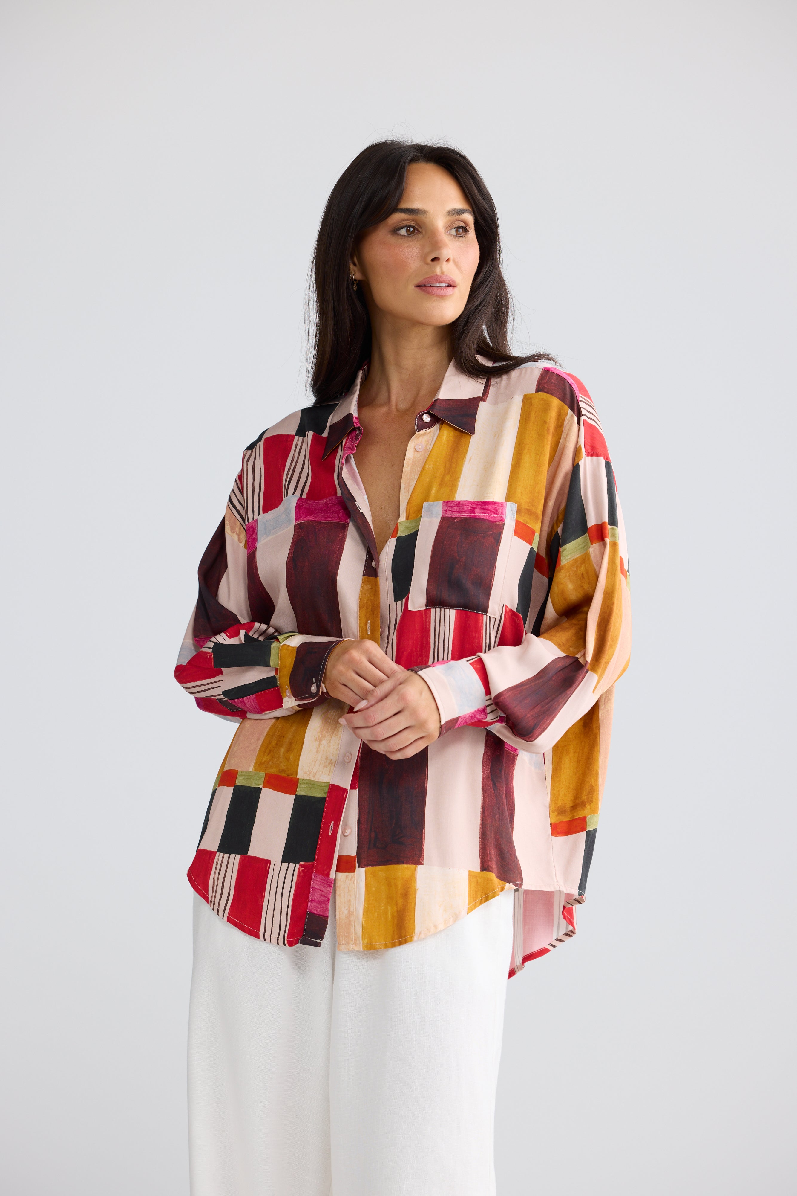 Image of A woman modeling a Brave & True long-sleeve button-up shirt with a colorful geometric patchwork pattern in red, yellow, pink, brown, and white, paired with white trousers, against a light gray studio background.