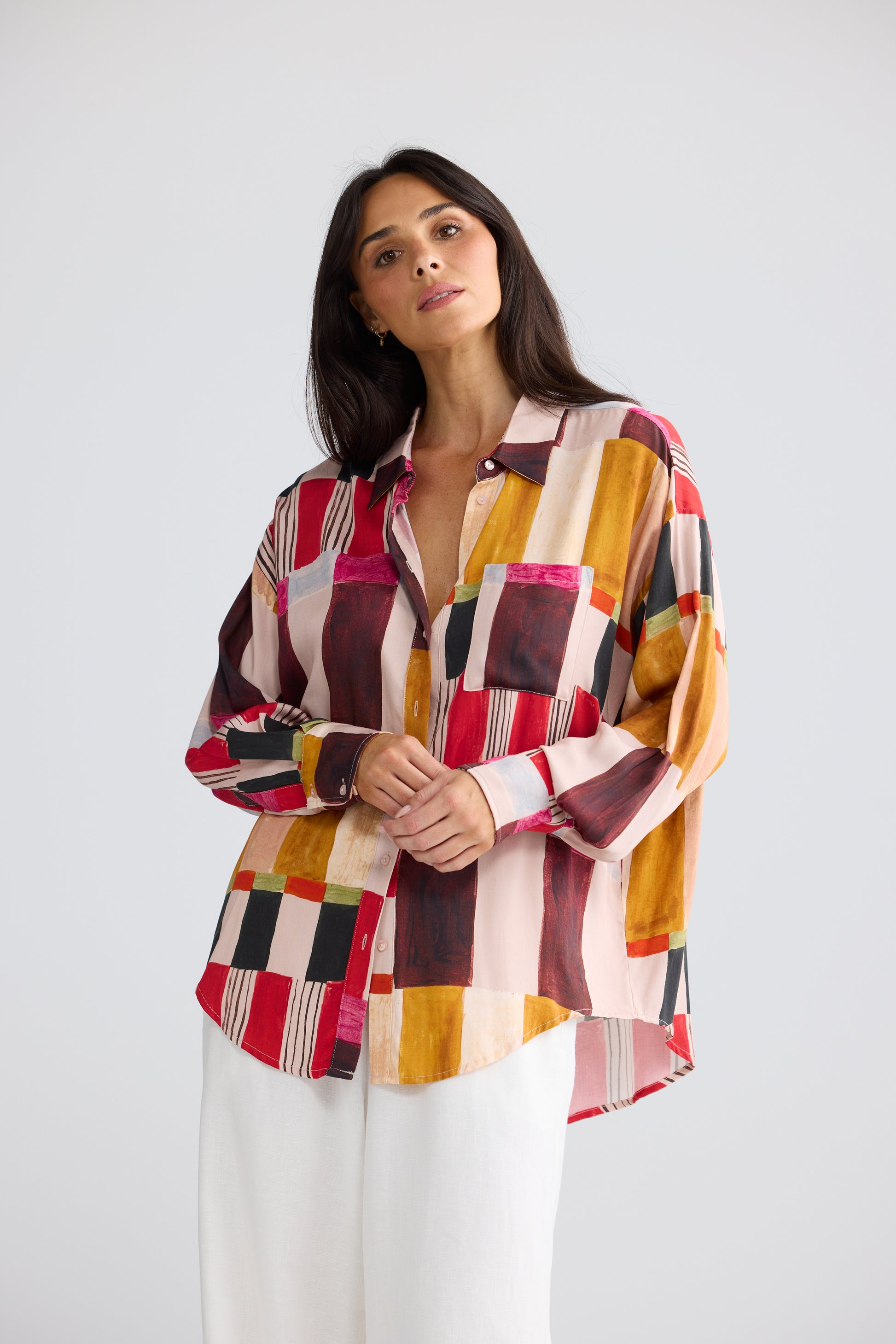 A woman modeling a Brave & True long-sleeve button-up shirt with a colorful geometric patchwork pattern in red, yellow, brown, black, and white, paired with white trousers, against a light gray studio background.