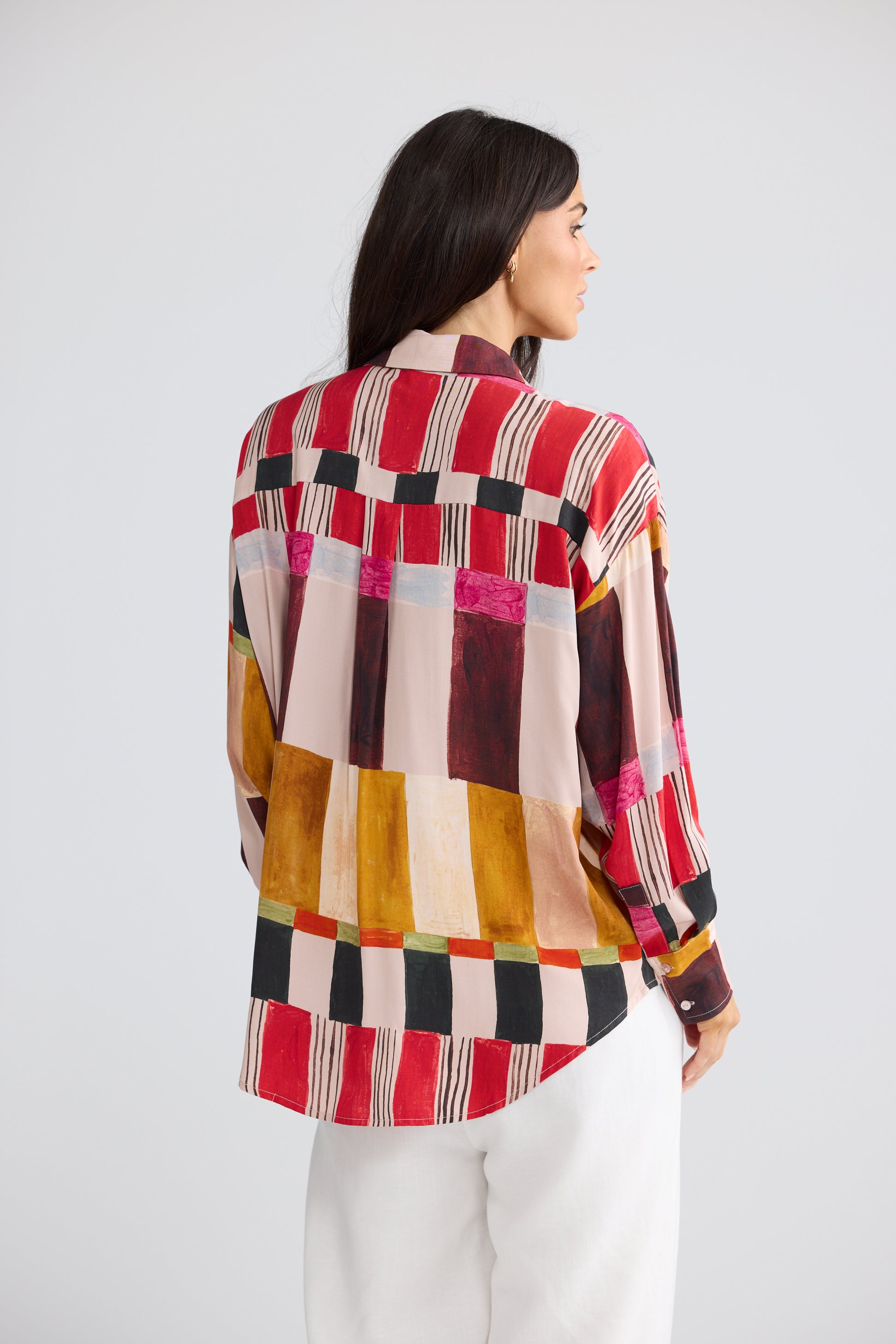 A woman shown from the back wearing a Brave & True long-sleeve patchwork button-up shirt with a multicolor geometric pattern in red, pink, yellow, black, and beige, paired with white pants, against a light gray studio background.