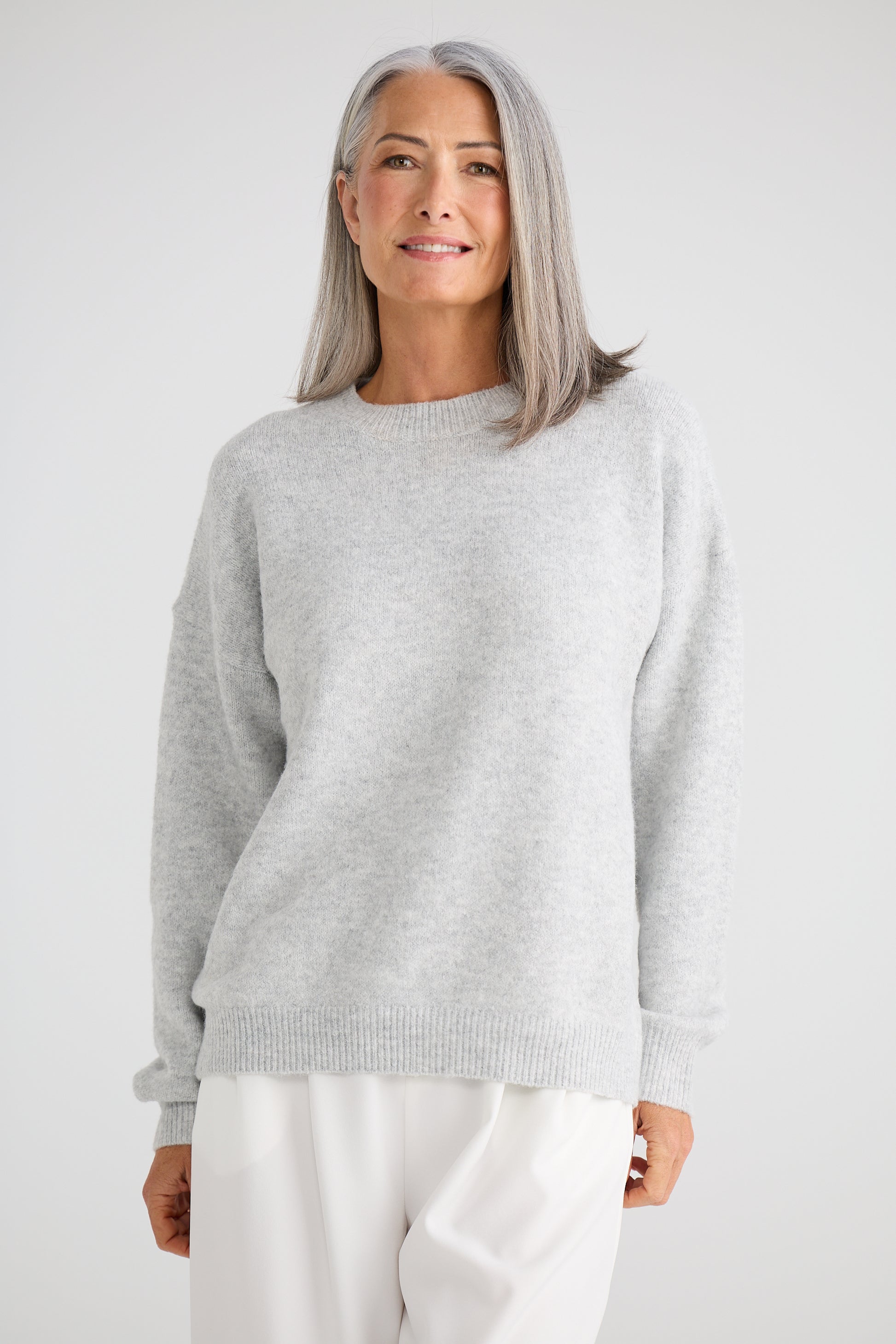 Woman wearing a Brave and True grey sweater and white pants against a plain background