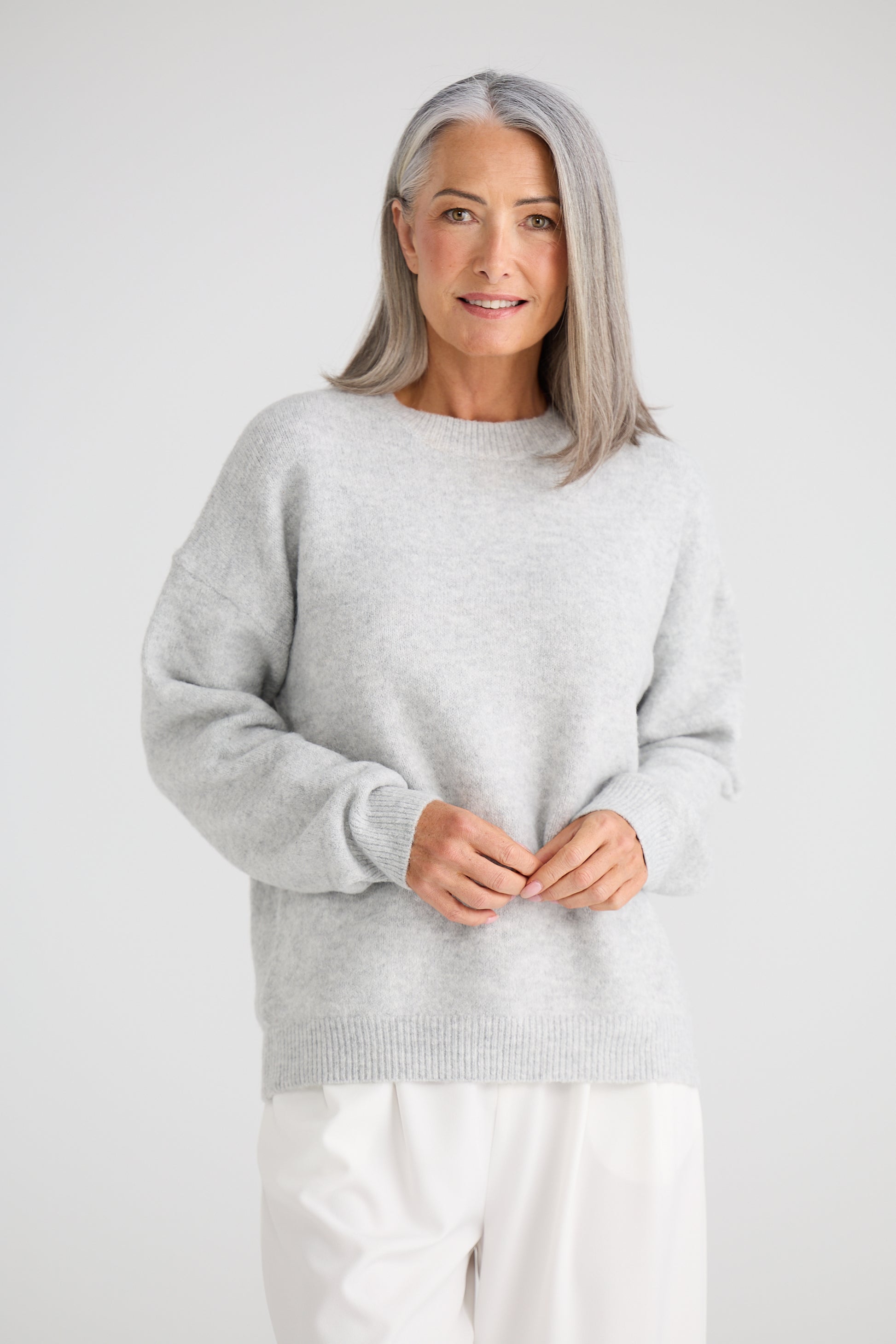 Woman wearing a Brave and True light grey sweater and white pants against a plain background