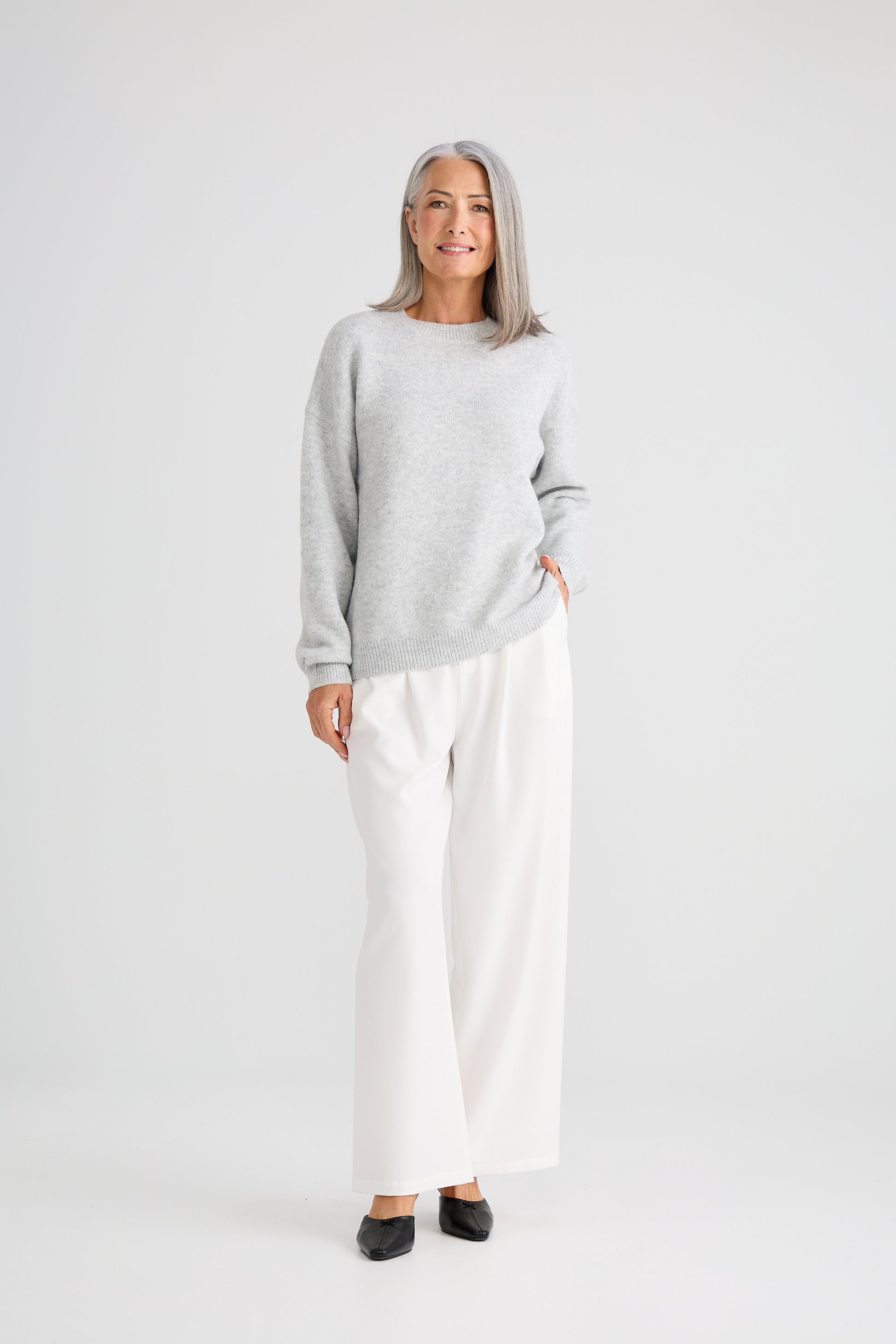 Woman wearing a Brave and True grey sweater and white pants on a white background