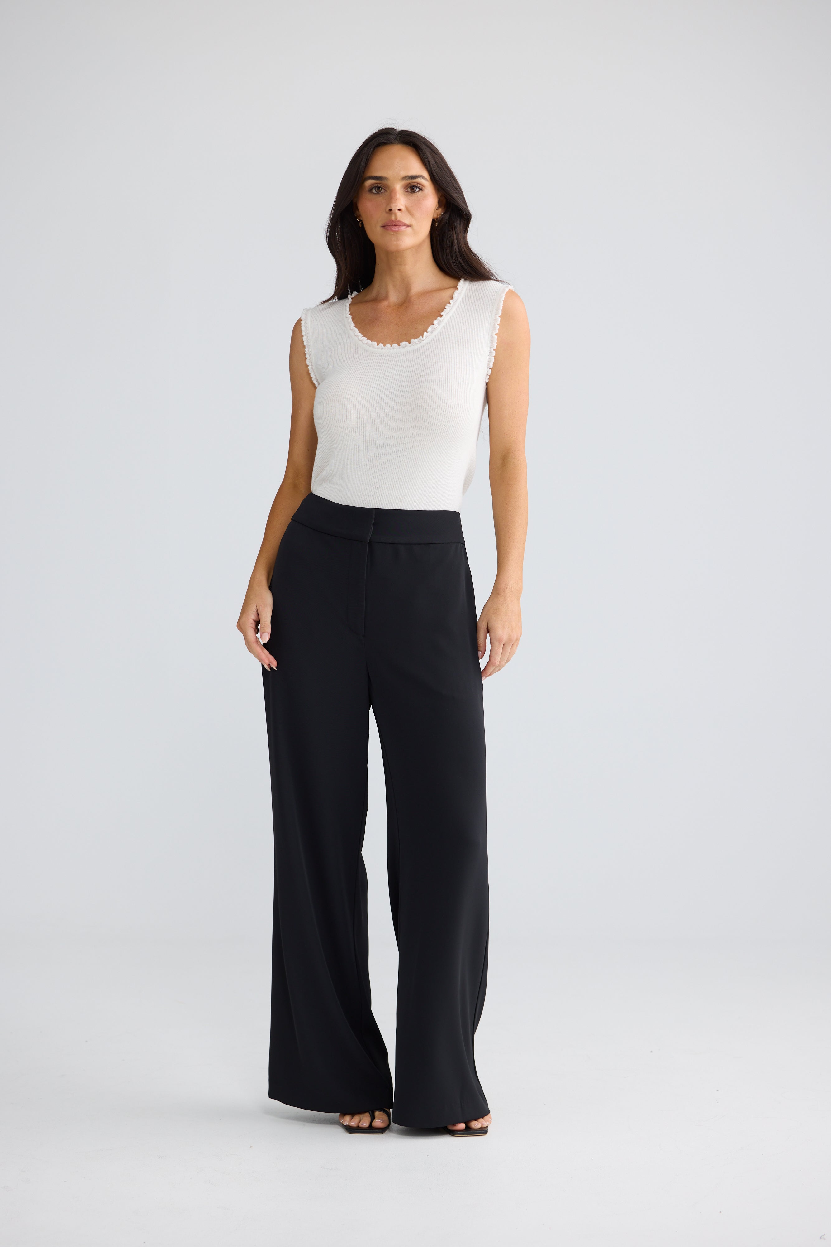 Image of A woman modeling a sleeveless white top with a scalloped neckline and Brave & True black wide-leg pants, standing against a light gray studio background.