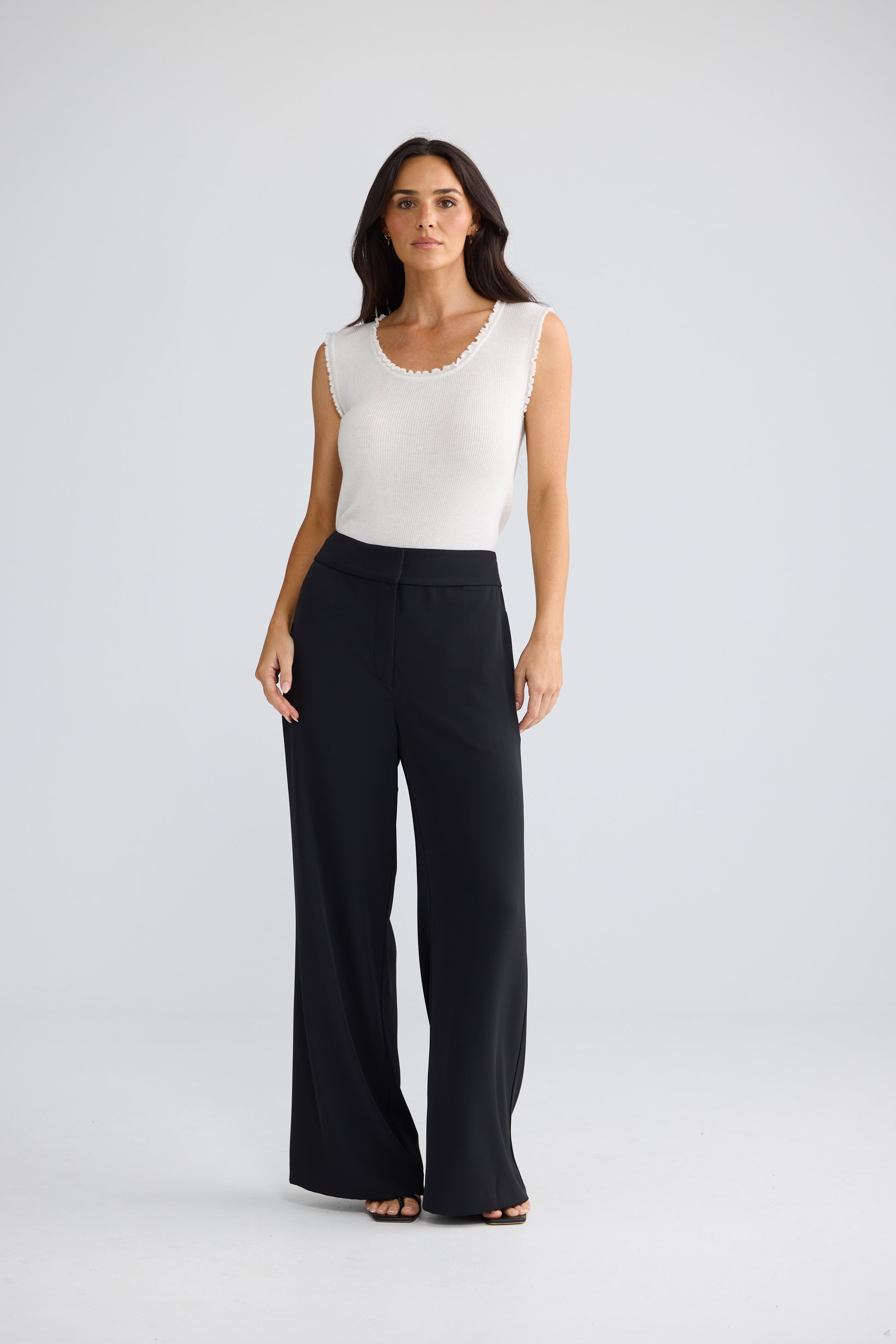A woman modeling a sleeveless white top with a scalloped neckline and Brave & True black wide-leg pants, standing against a light gray studio background.