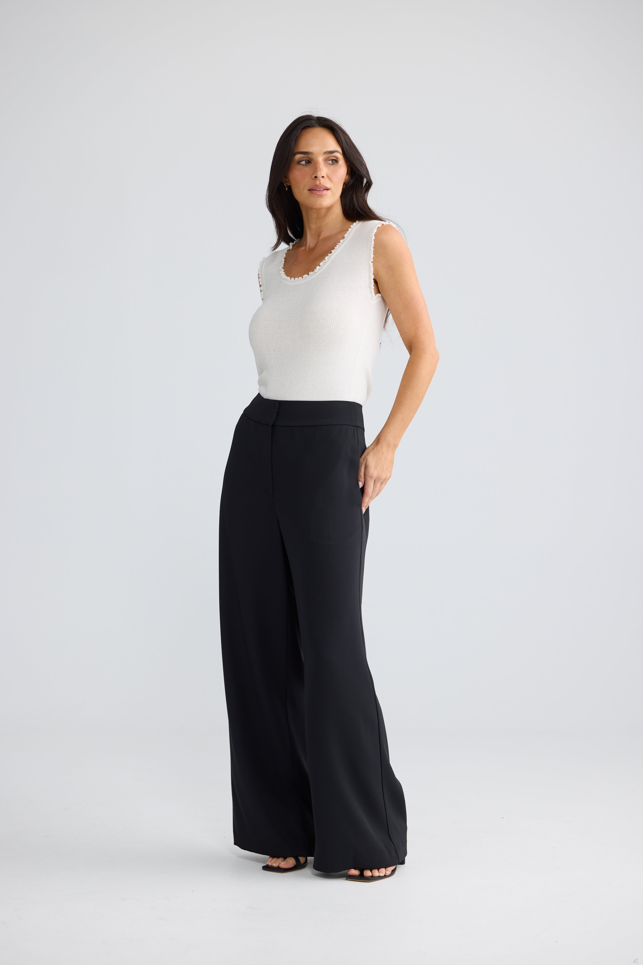 Image of A woman modeling a pair of Brave & True black wide-leg trousers with a white sleeveless top, standing against a light gray studio background.