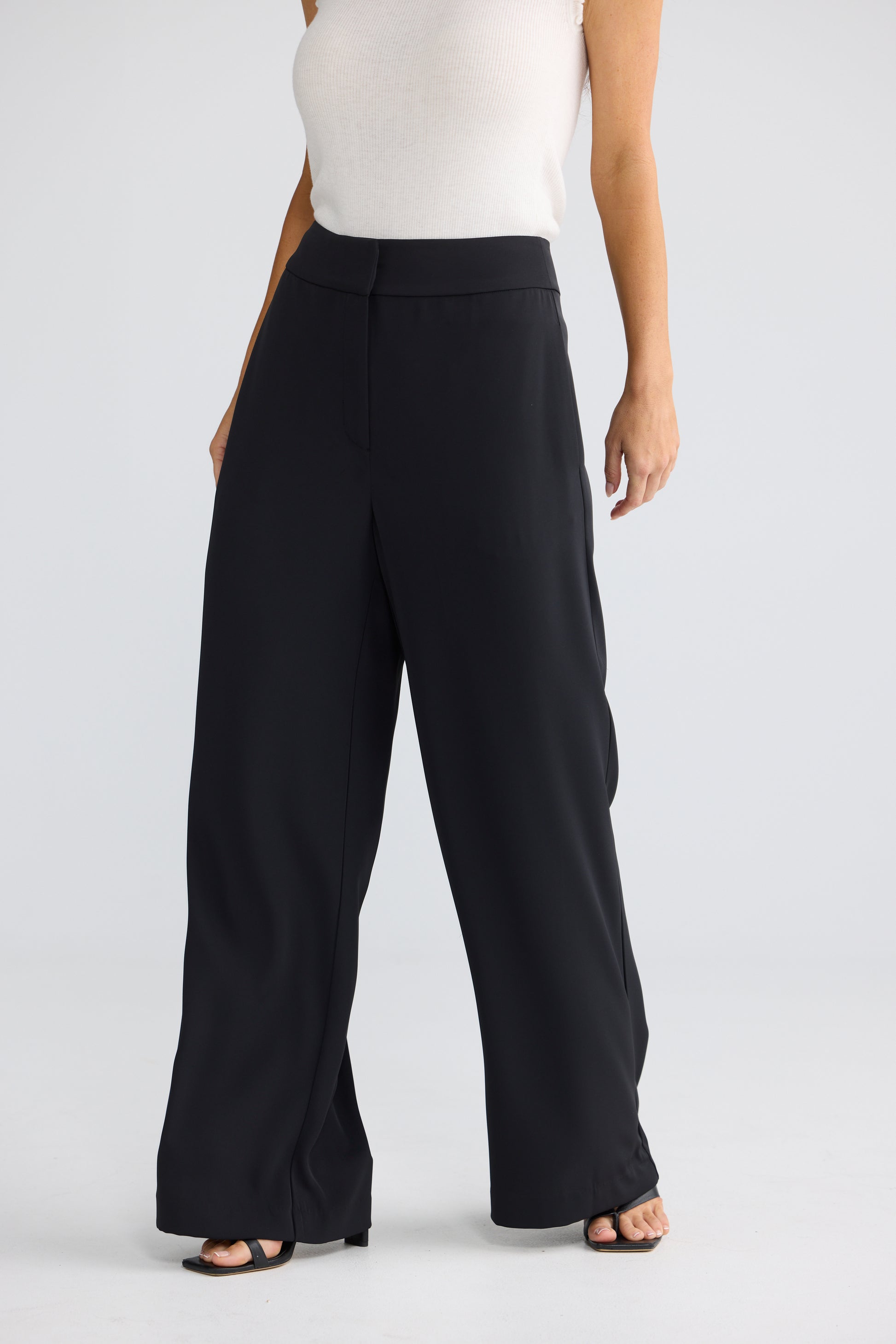 A model wearing Brave & True black wide-leg cropped trousers with a high waist, paired with a white sleeveless top and black sandals, shown from the waist down on a light gray studio background.