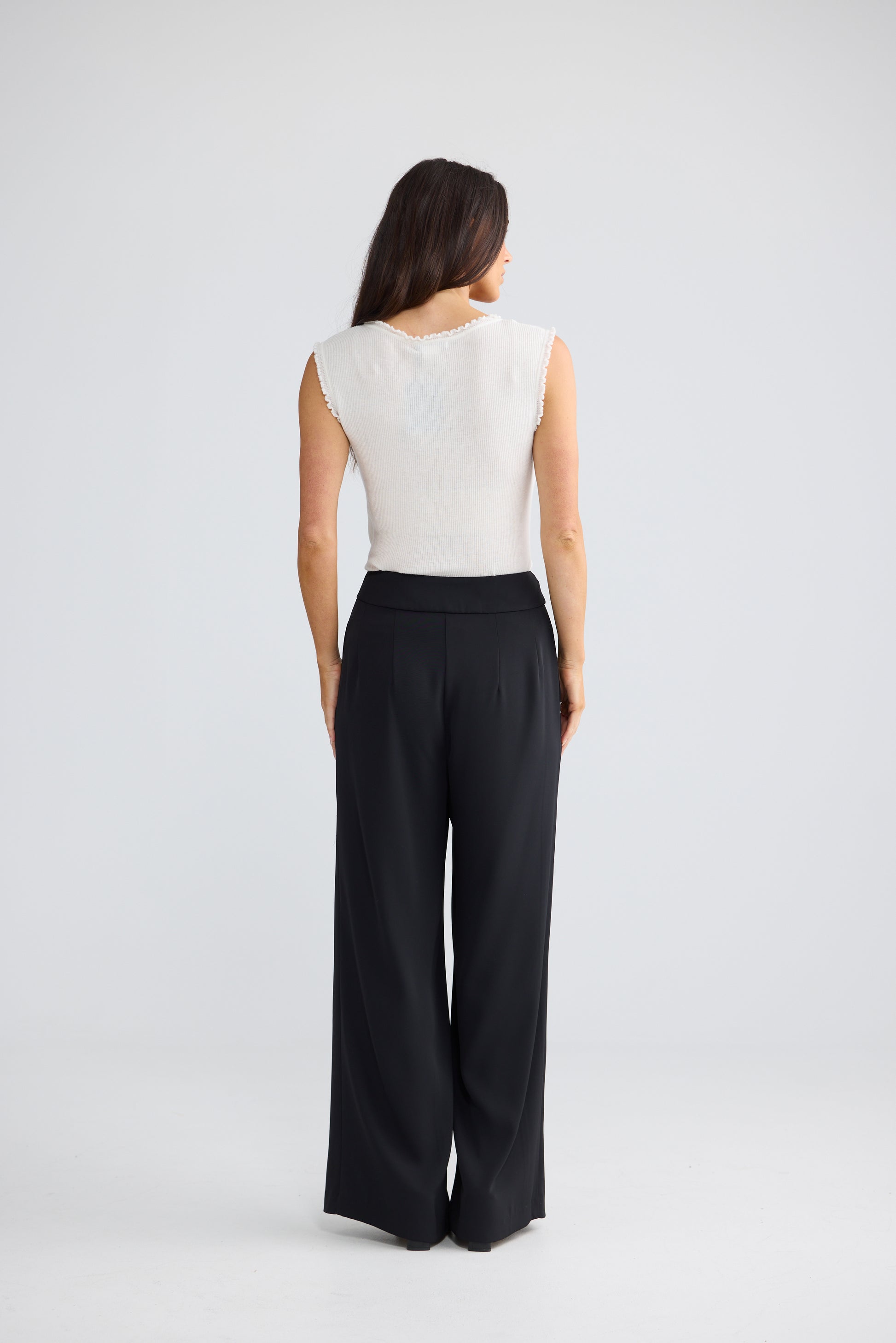 A woman shown from the back wearing a sleeveless white top and Brave & True black wide-leg trousers, standing against a light gray studio background.