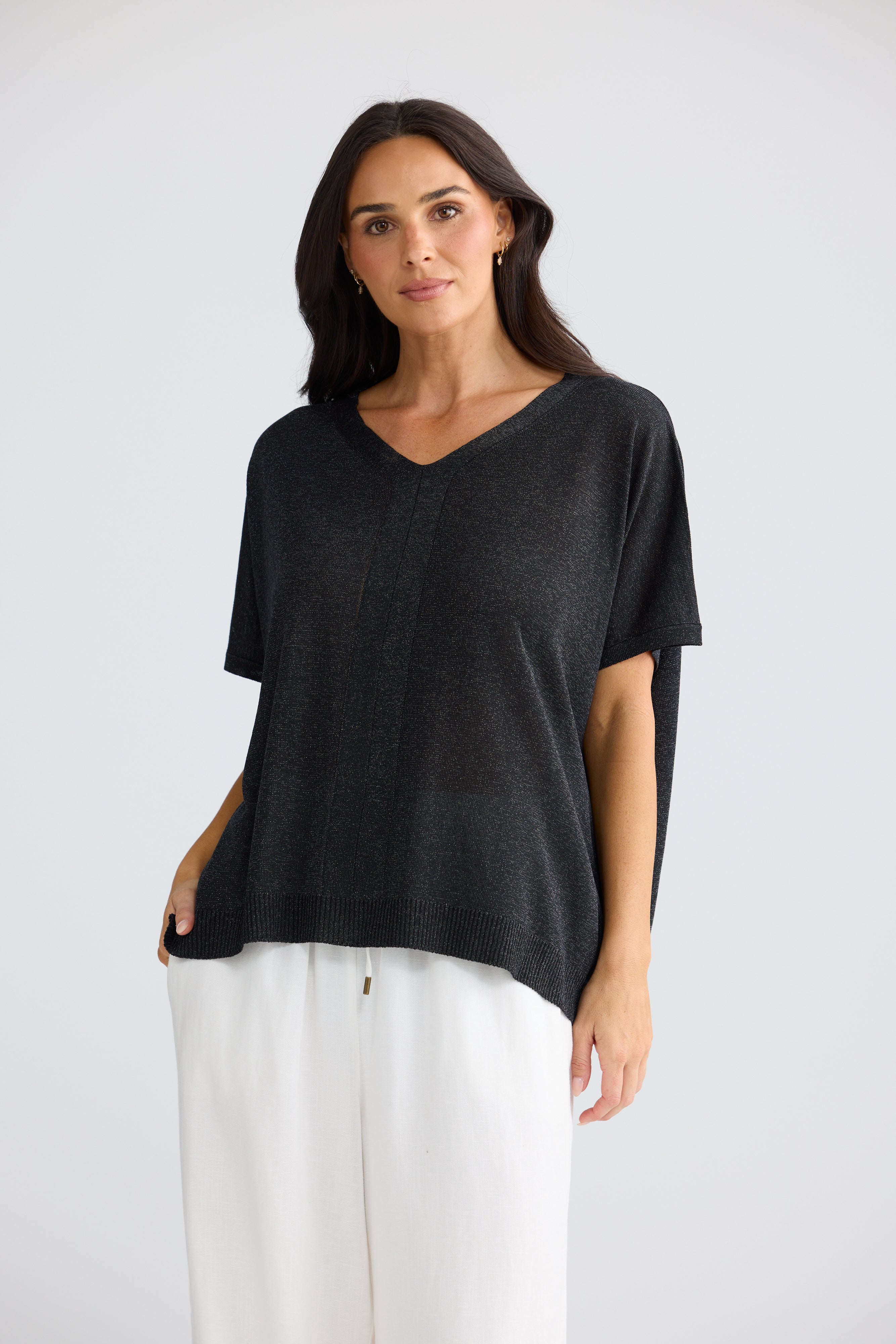 Image of A woman modeling a Brave & True black short-sleeve knit top with a V-neck and relaxed fit, paired with white pants, against a light gray studio background.