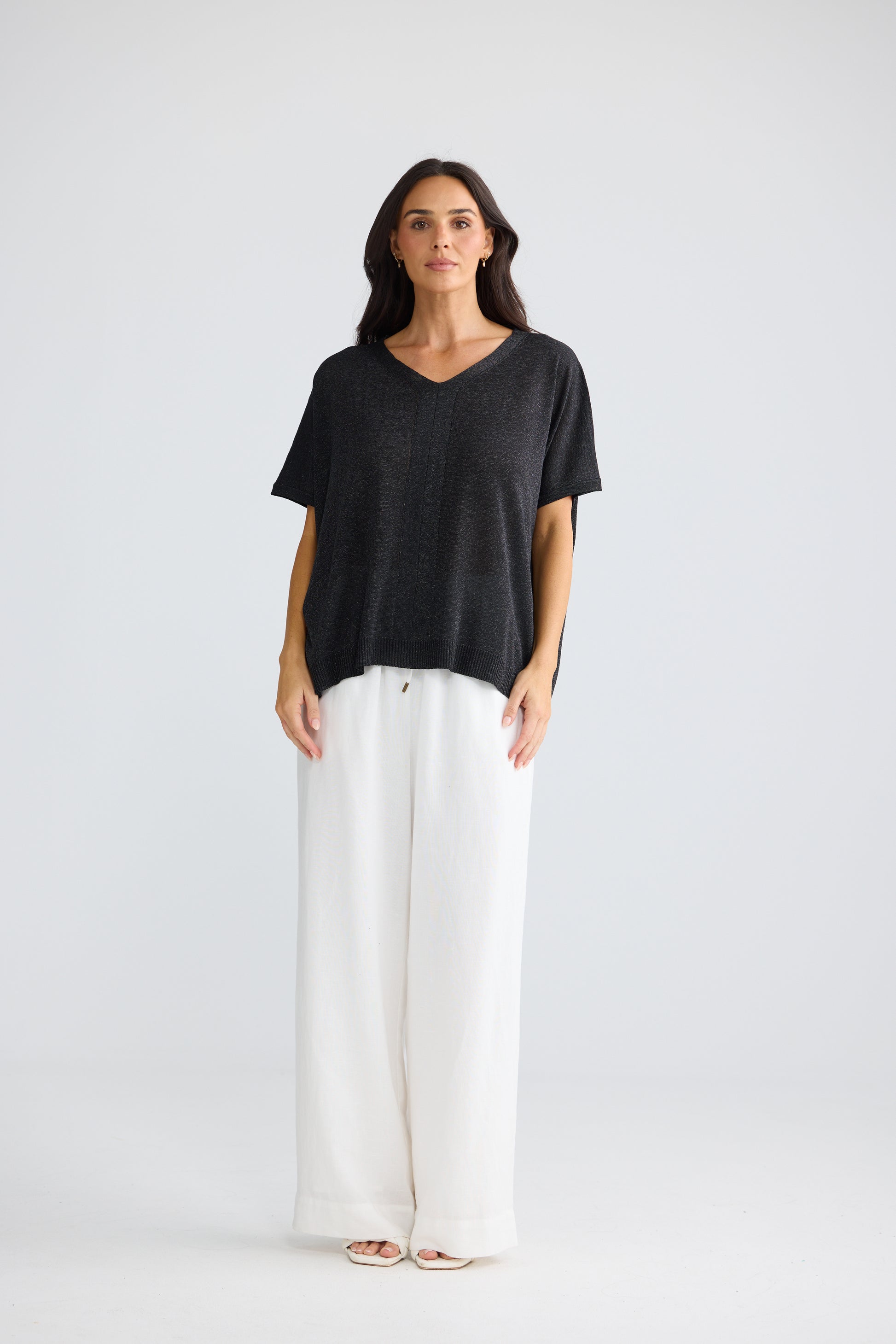 A woman stands against a light gray studio background wearing a Brave & True loose black short-sleeve V-neck top and wide-leg white pants.
