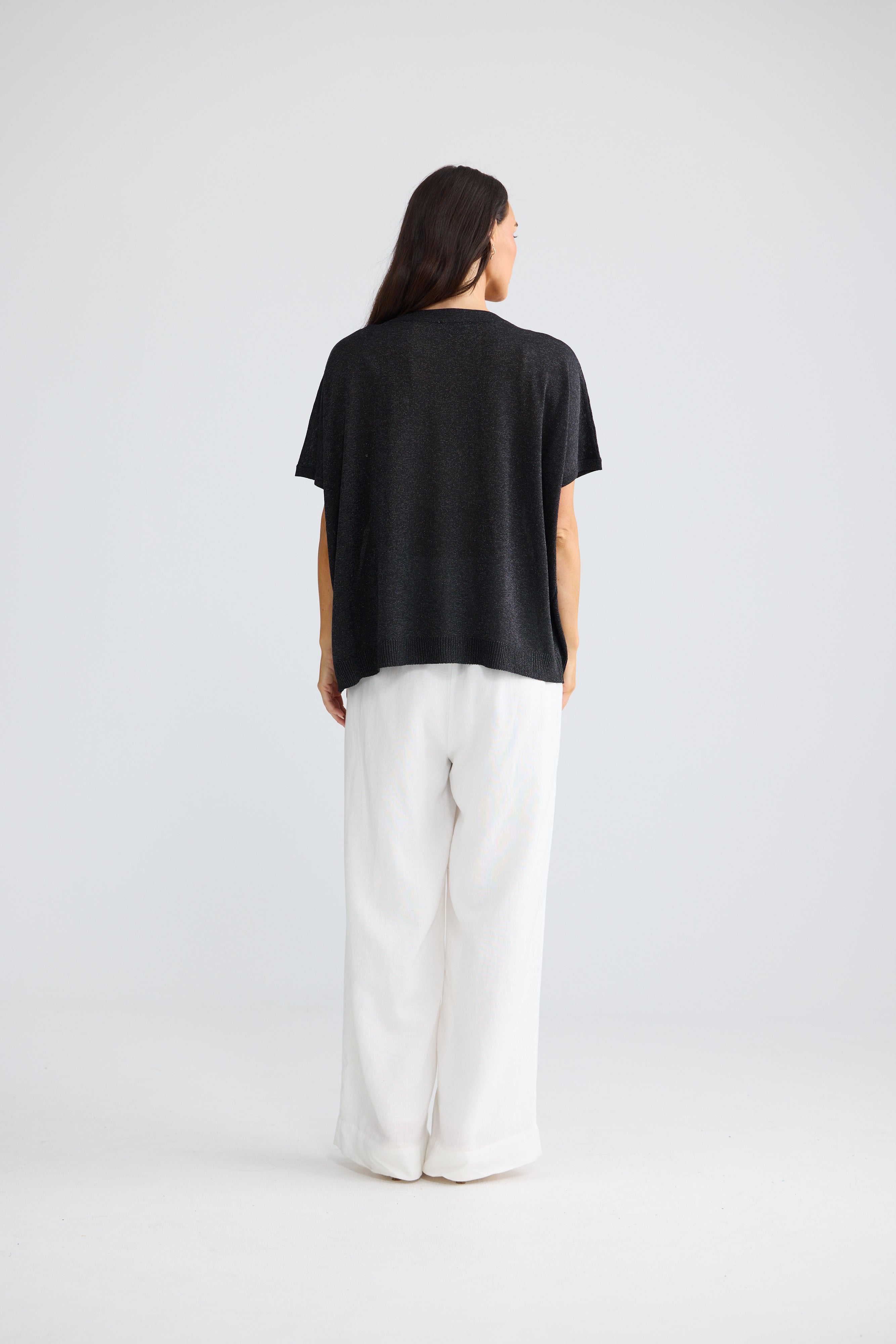 Image of A woman is shown from behind wearing a Brave & True black short-sleeve top and white wide-leg pants against a light gray studio background.
