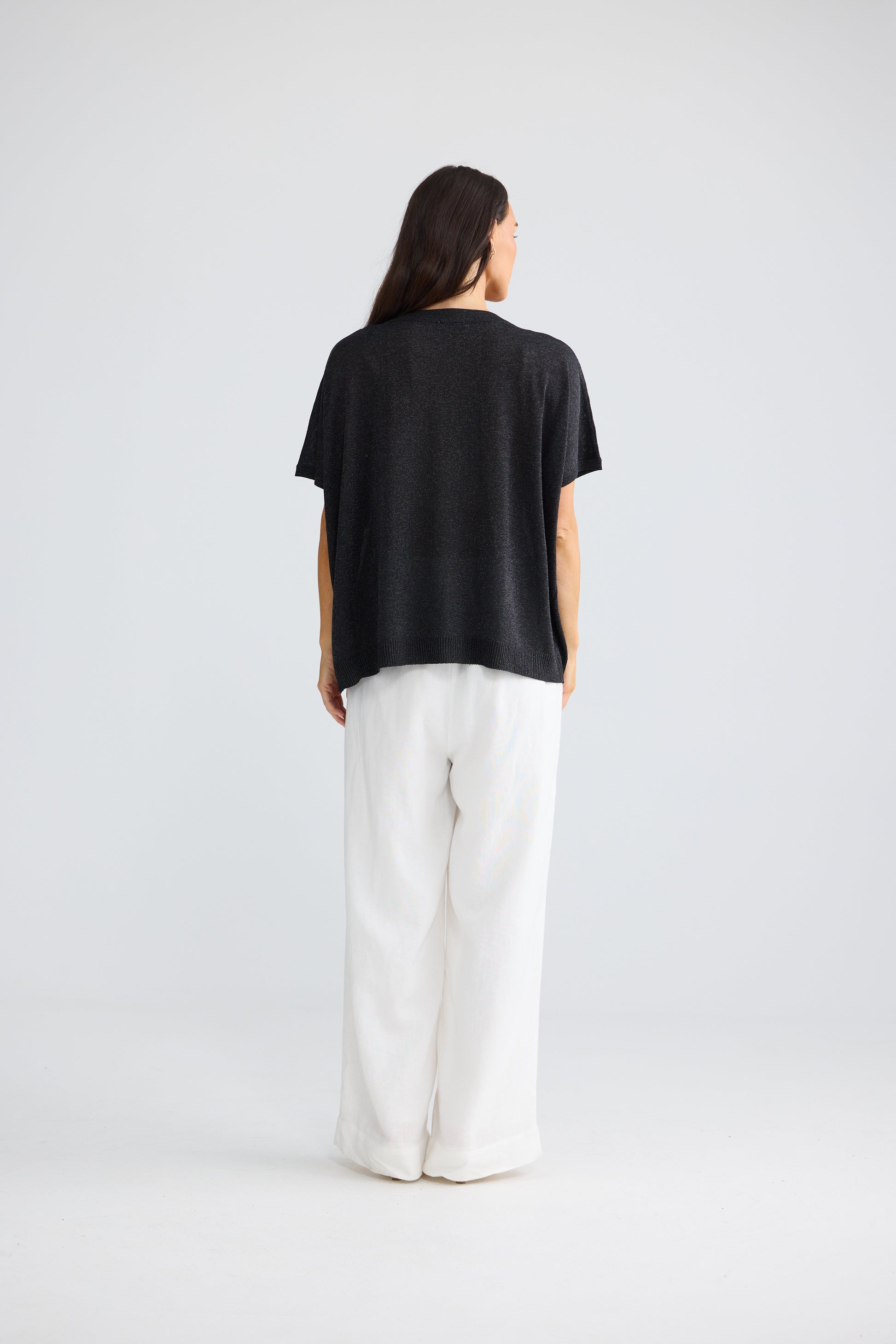 A woman is shown from behind wearing a Brave & True black short-sleeve top and white wide-leg pants against a light gray studio background.