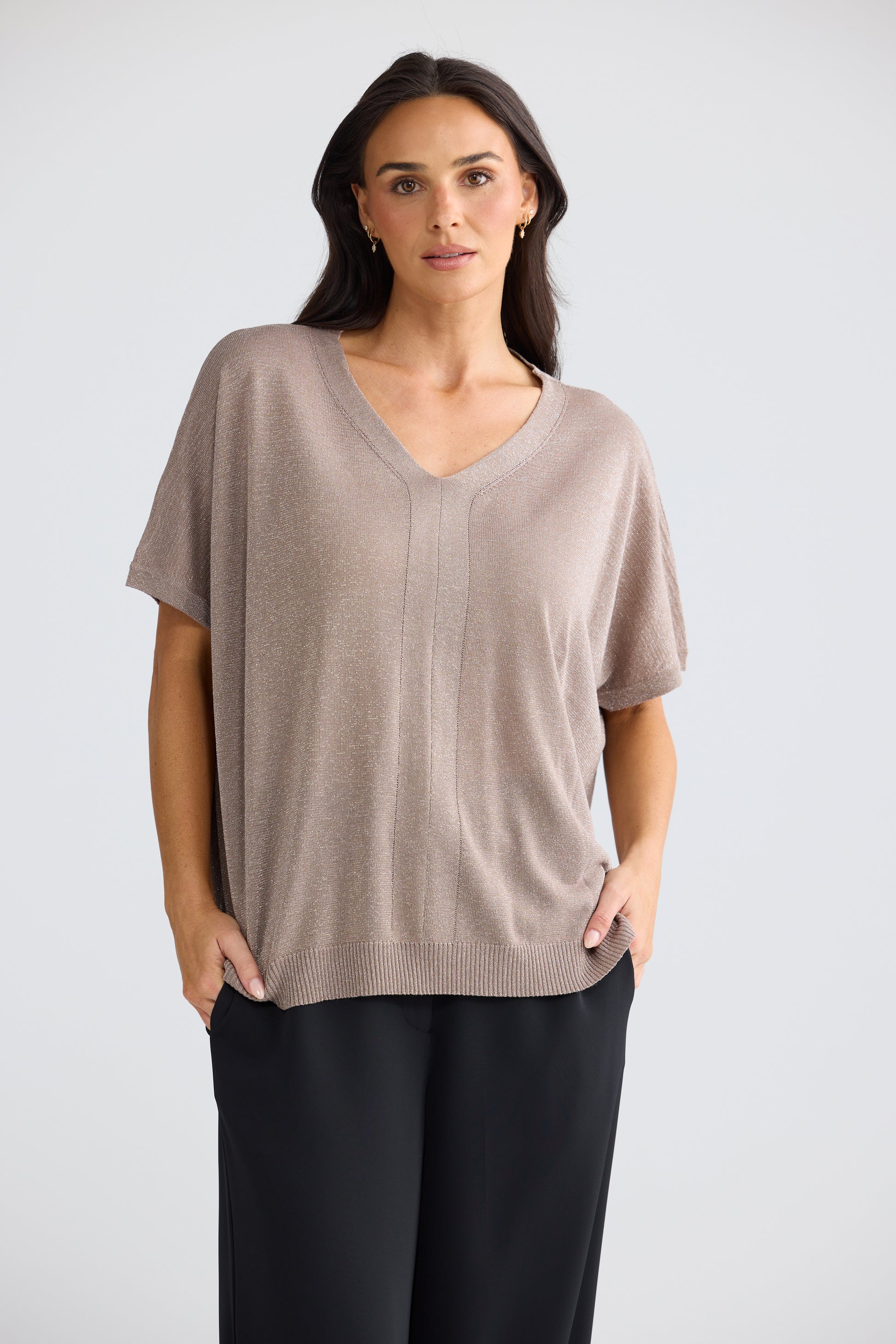 A woman modeling a Brave & True short-sleeve knit V-neck top in a light taupe-beige color, paired with black pants, against a plain light gray studio background.