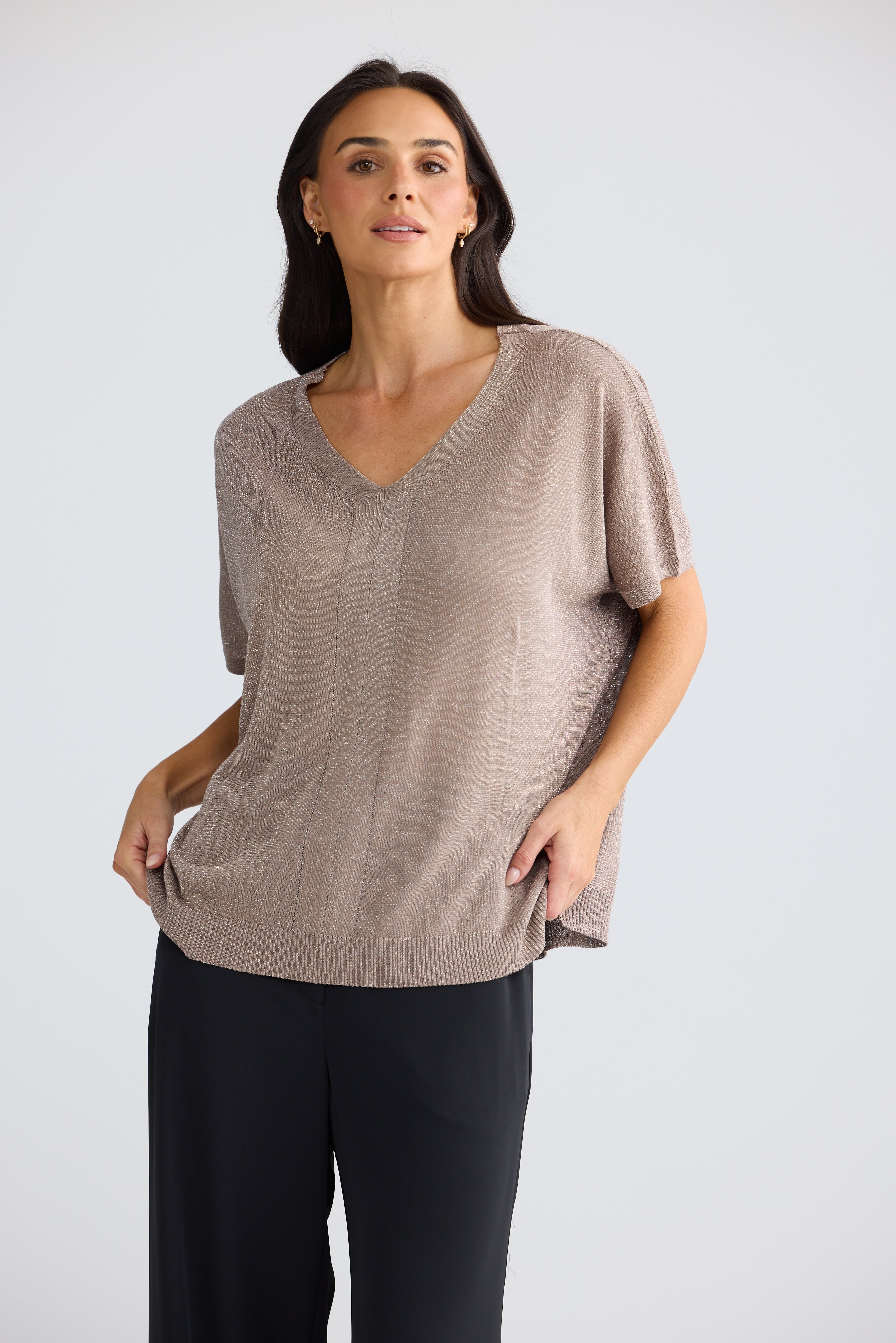 Image of A woman modeling a Brave & True short-sleeve V-neck knit top in taupe, paired with black pants, against a light gray studio background.