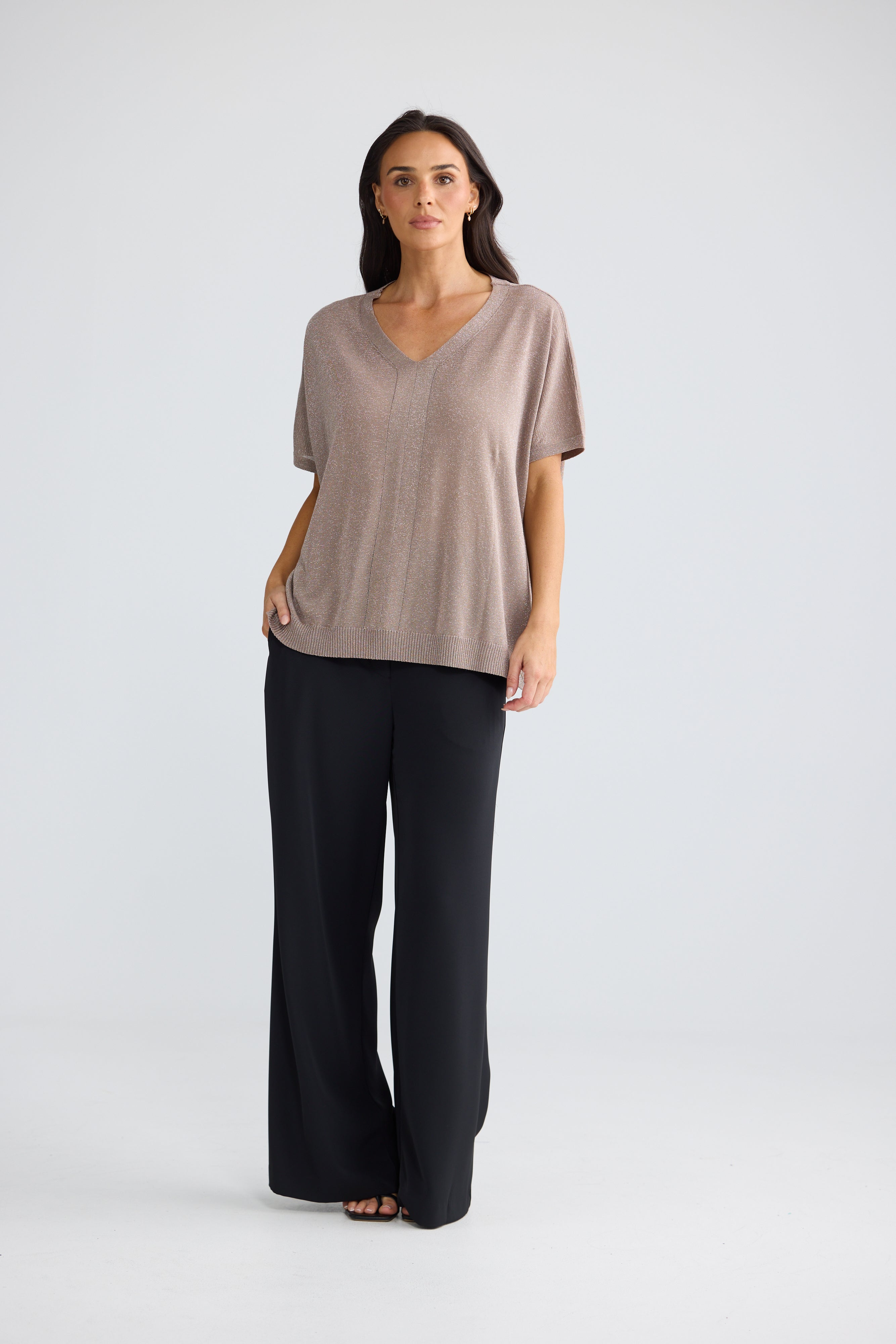 Image of A woman modeling a Brave & True short-sleeve V-neck top in a muted taupe-beige color, paired with black wide-leg pants, against a light gray studio background.