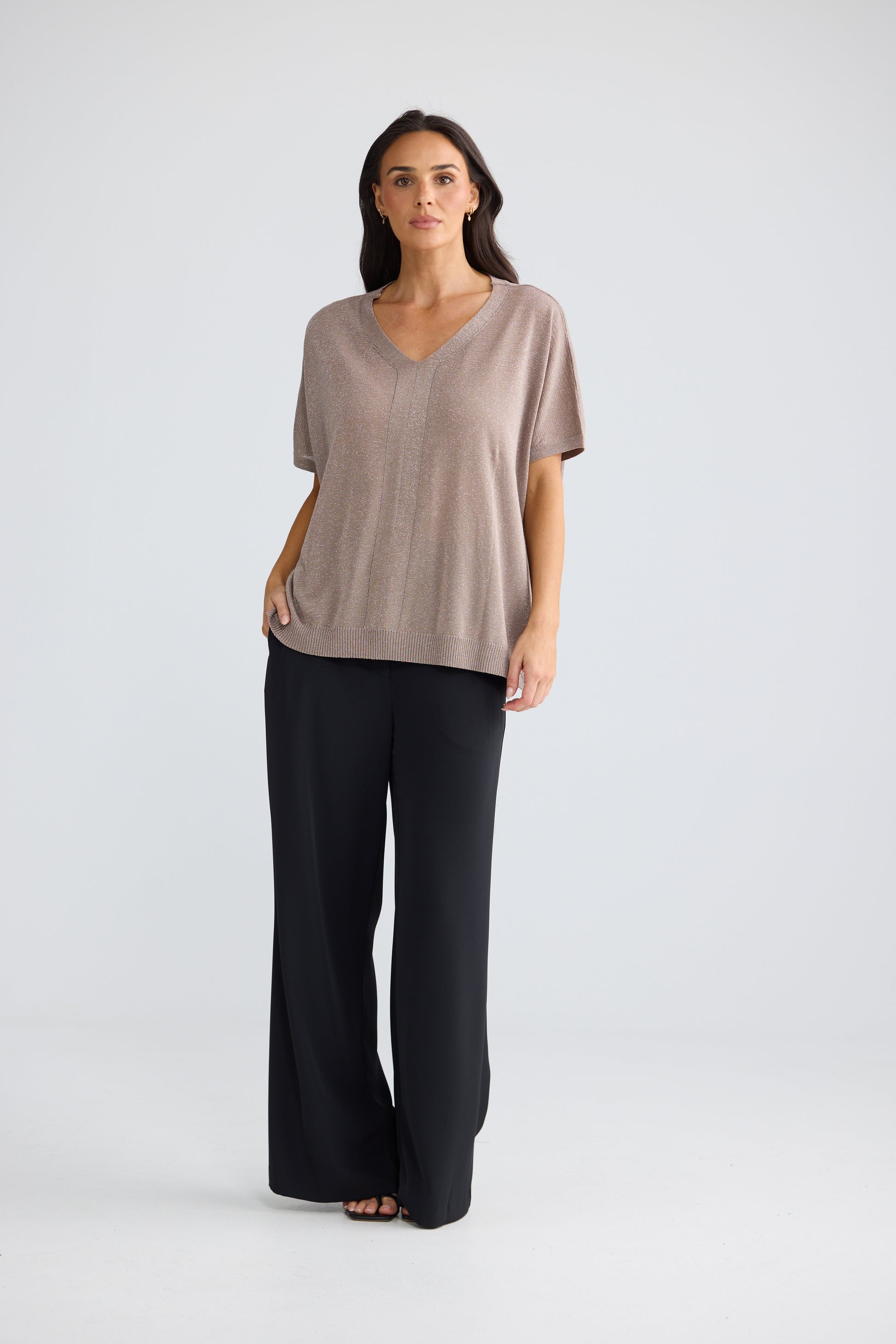 A woman modeling a Brave & True short-sleeve V-neck top in a muted taupe-beige color, paired with black wide-leg pants, against a light gray studio background.