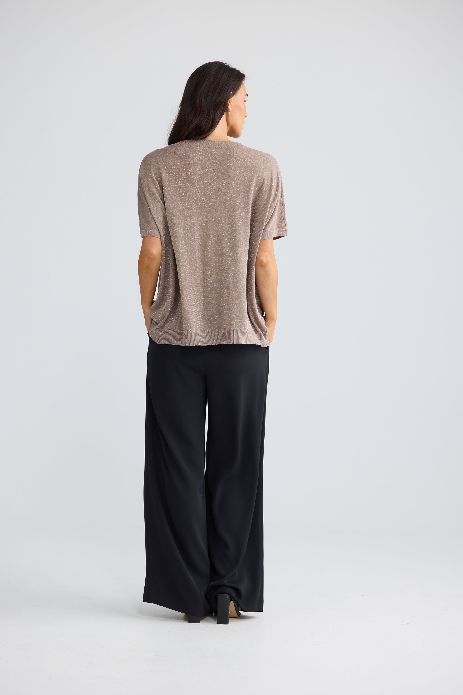 A woman is shown from the back wearing a Brave & True short-sleeve taupe knit top and black wide-leg trousers against a light gray studio background.
