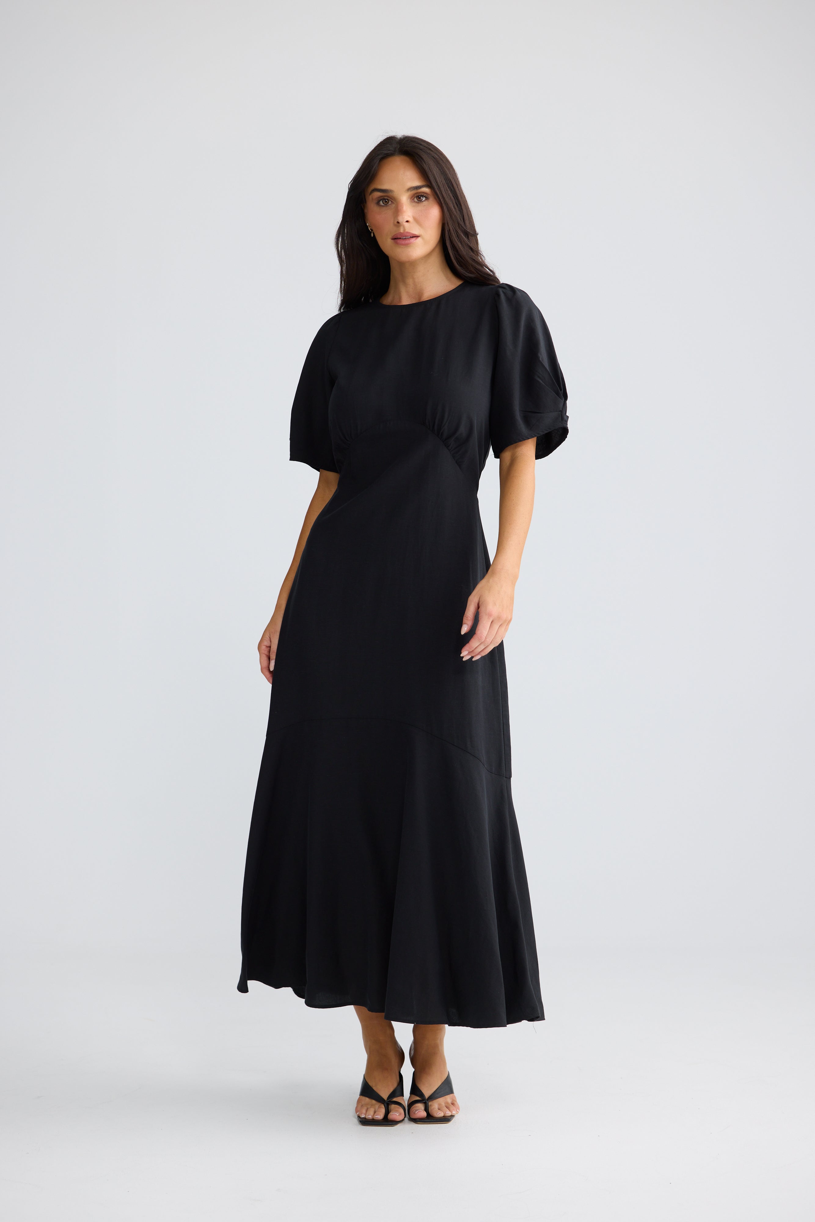 Image of A woman modeling a Brave & True black midi dress with short puff sleeves, a boat neckline, and a gathered waist detail, photographed against a light gray studio background.