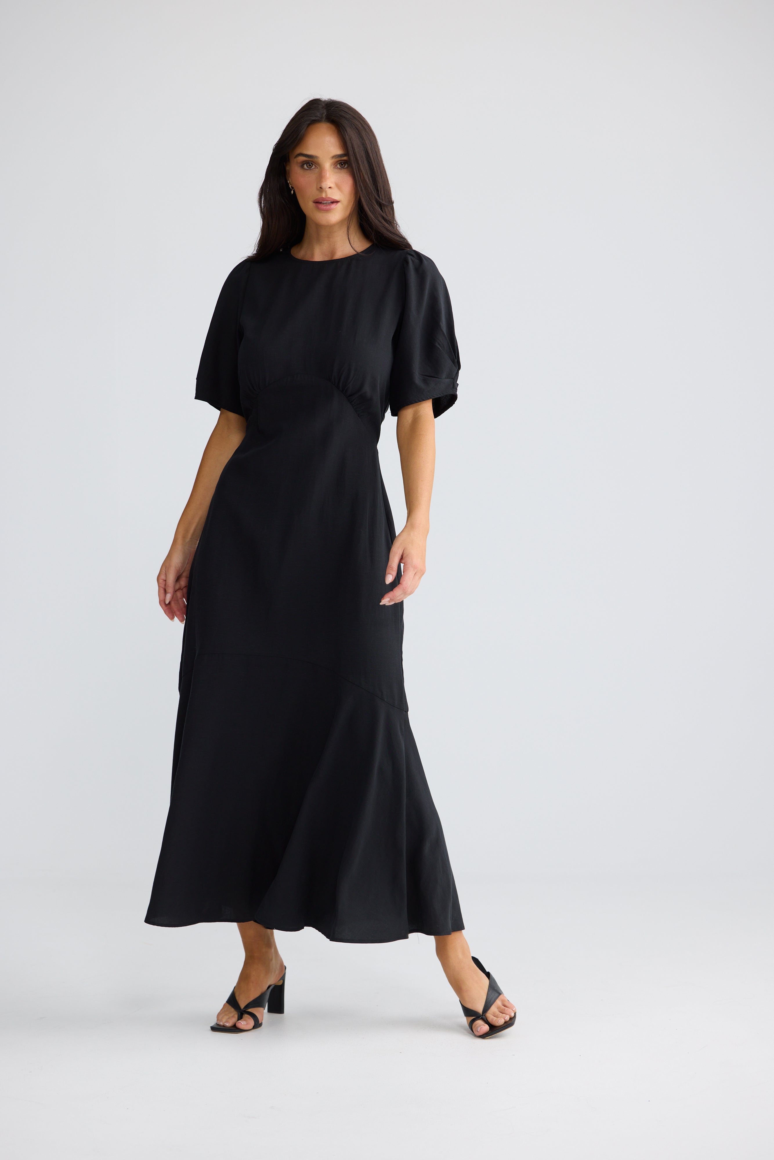 Image of A woman modeling a Brave & True black midi dress with short puff sleeves, a round neckline, and a flared skirt, photographed against a light gray studio background.