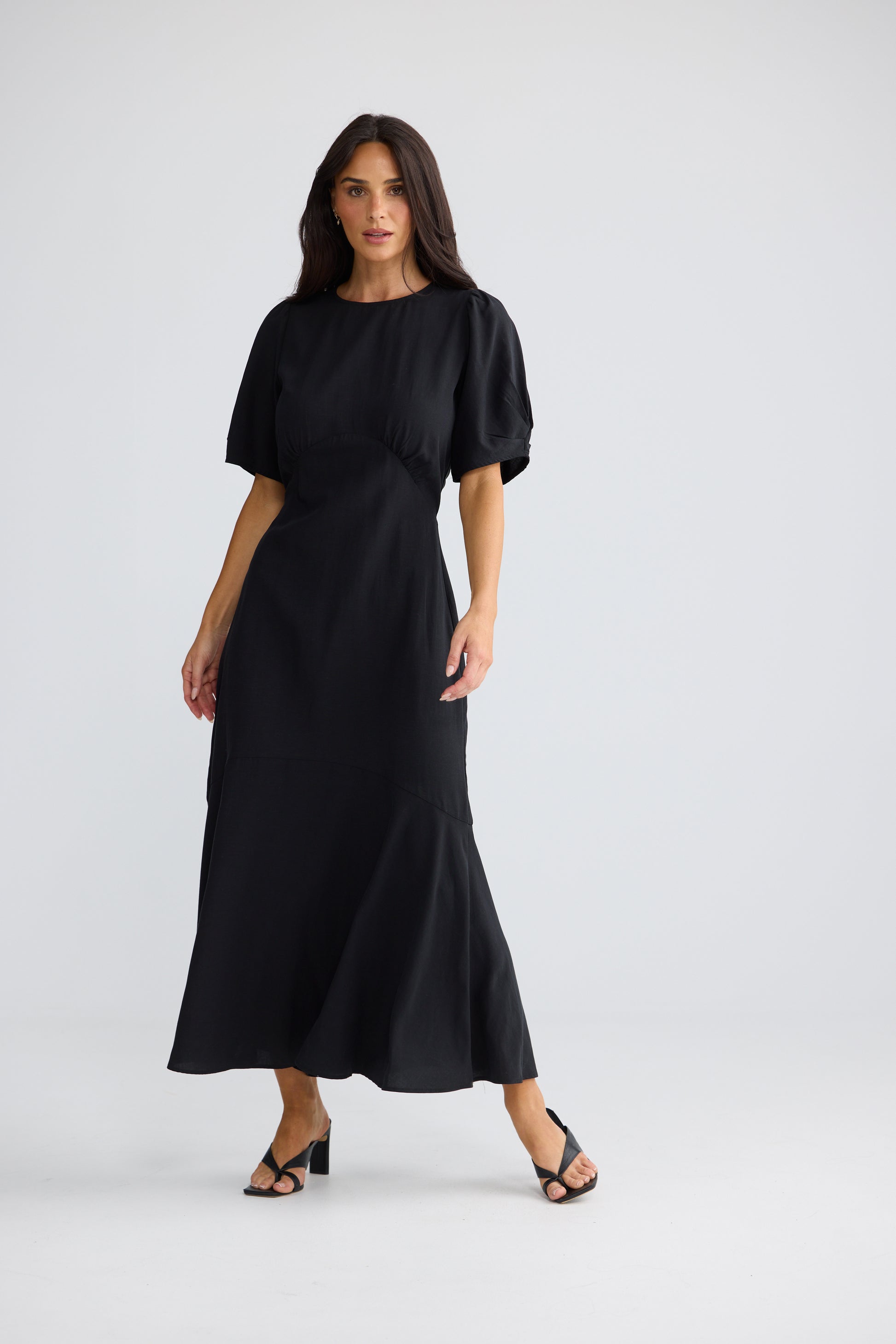 A woman modeling a Brave & True black midi dress with short puff sleeves, a round neckline, and a flared skirt, photographed against a light gray studio background.