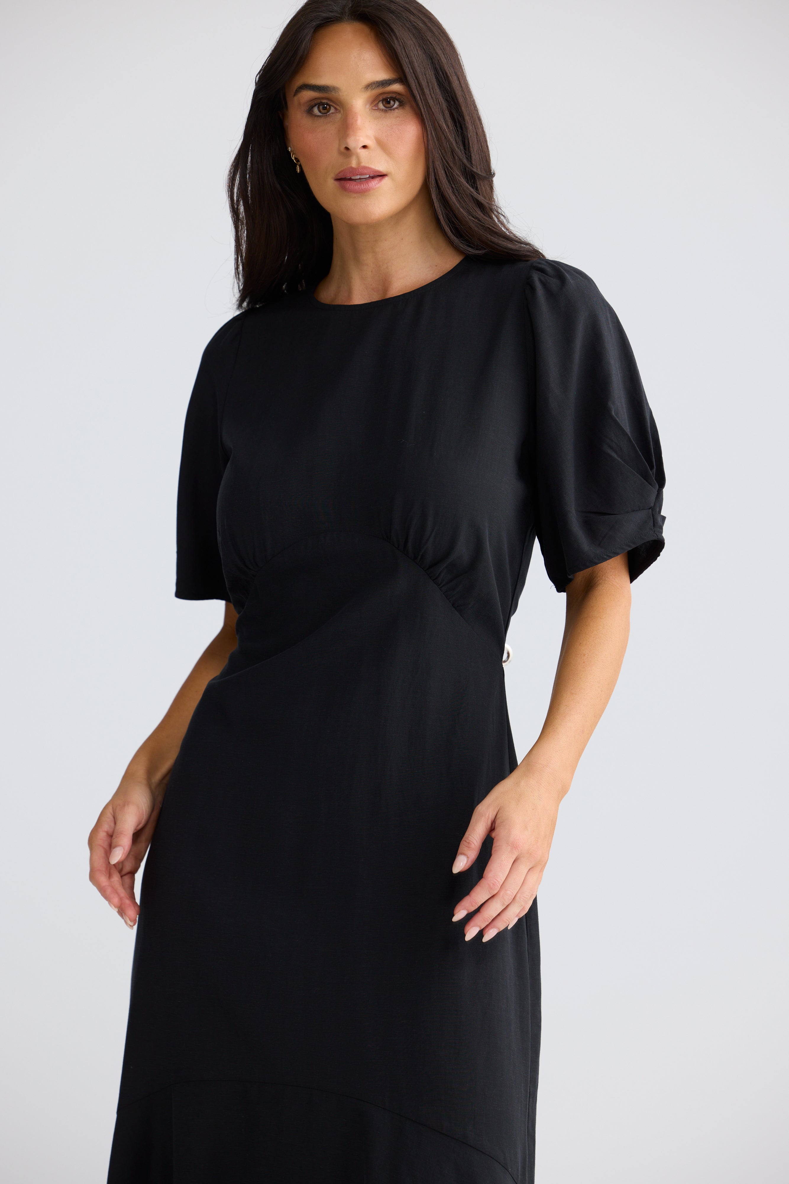 Image of A woman modeling a Brave & True black midi dress with short puff sleeves, a round neckline, and a gathered waist detail, photographed against a light gray studio background.