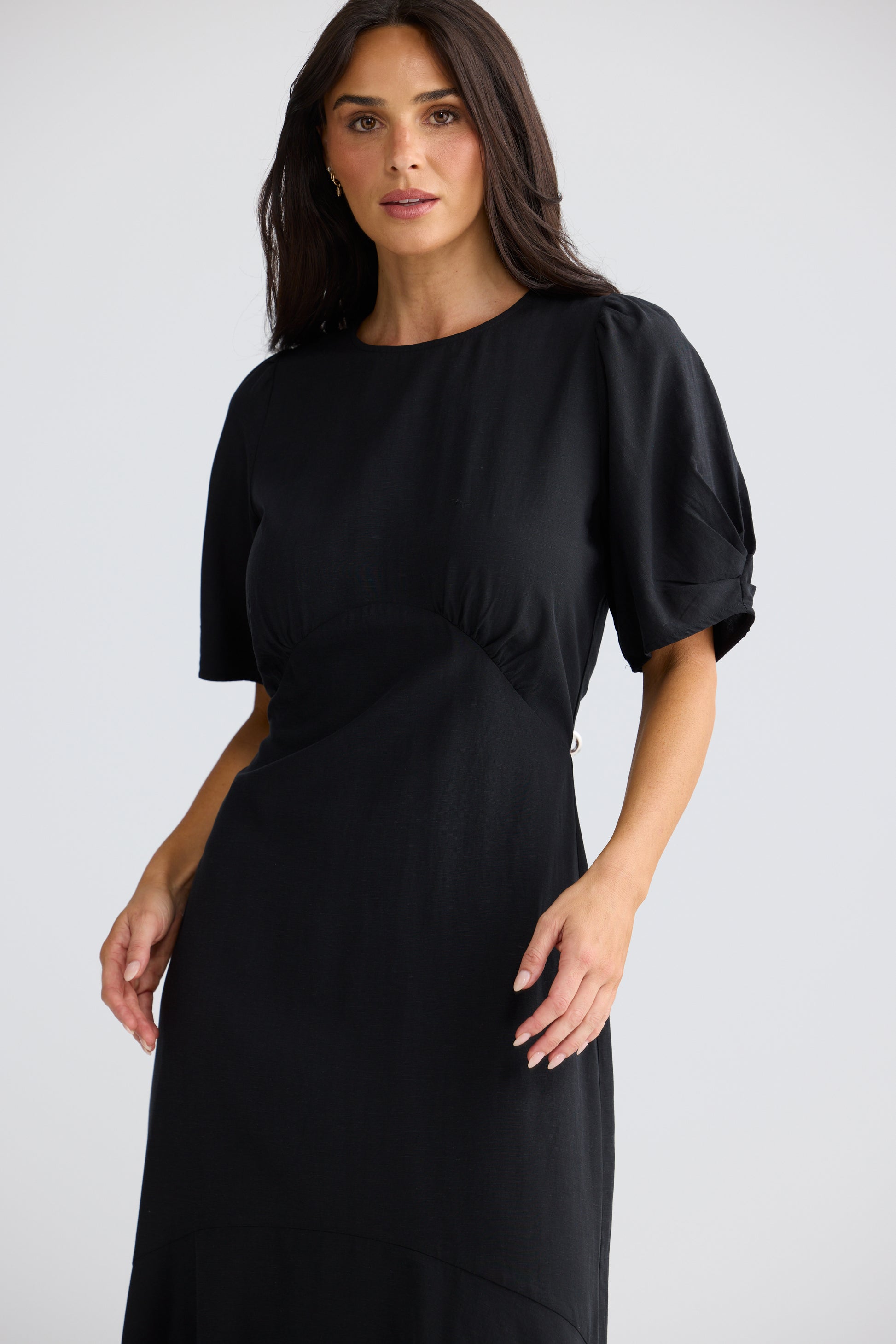 A woman modeling a Brave & True black midi dress with short puff sleeves, a round neckline, and a gathered waist detail, photographed against a light gray studio background.