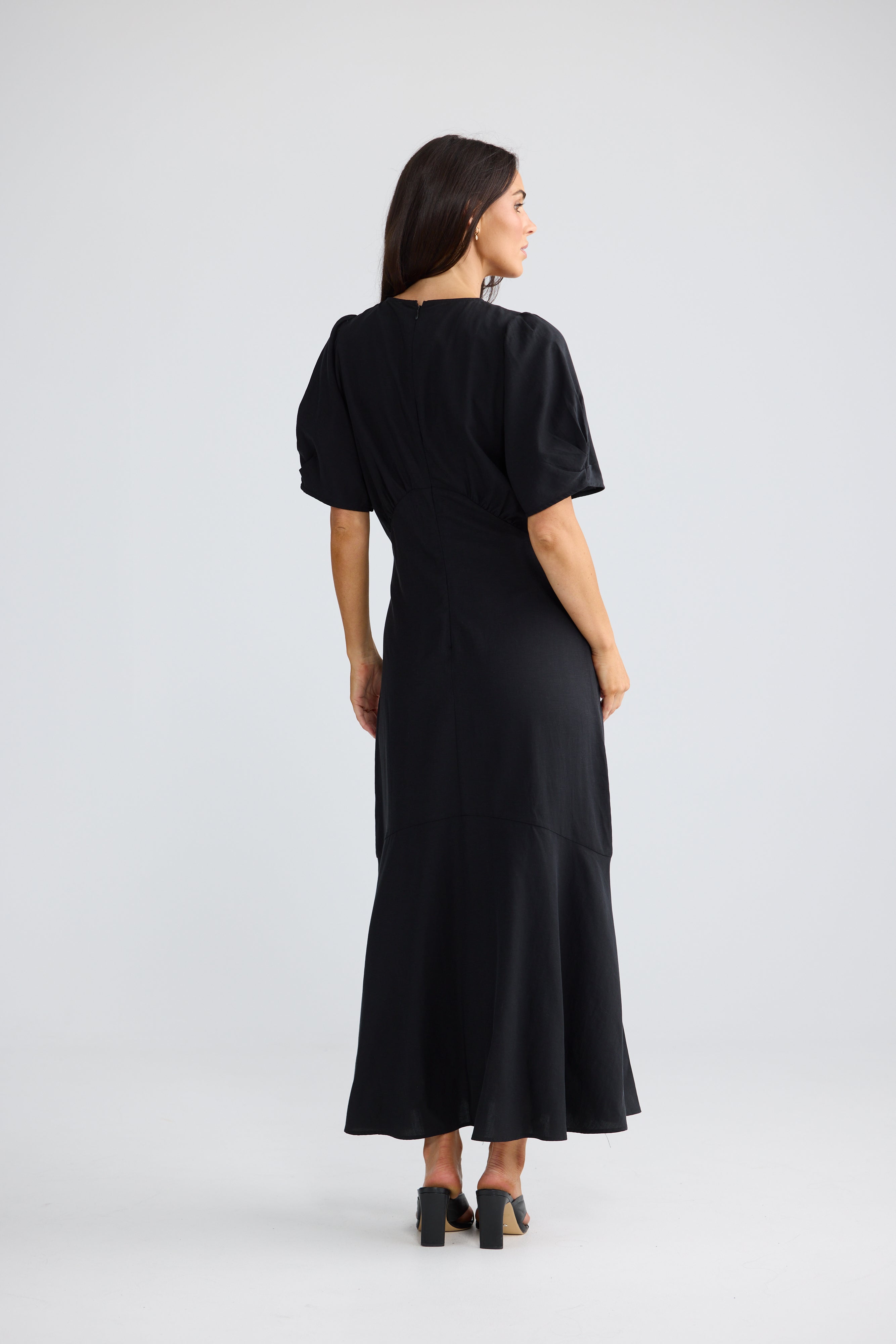 Image of A woman wearing a Brave & True black midi dress with short puff sleeves and a flared hem, shown from the back against a light gray studio background.