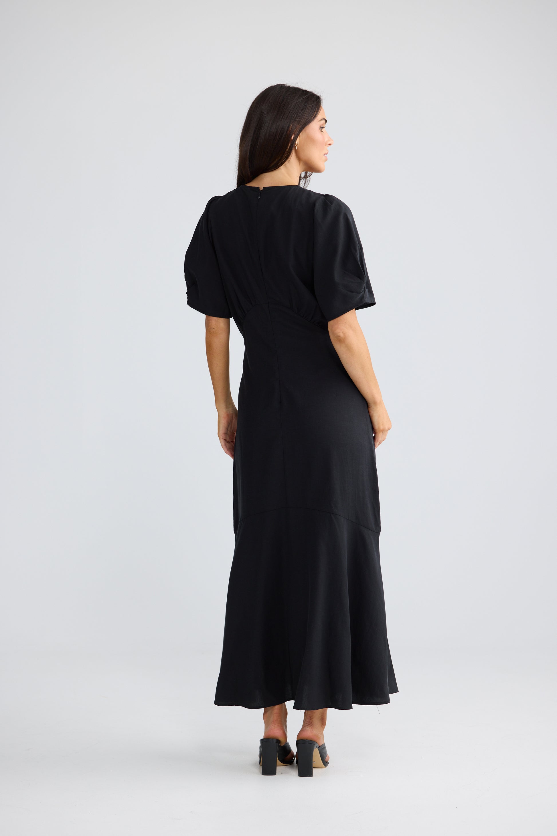 A woman wearing a Brave & True black midi dress with short puff sleeves and a flared hem, shown from the back against a light gray studio background.