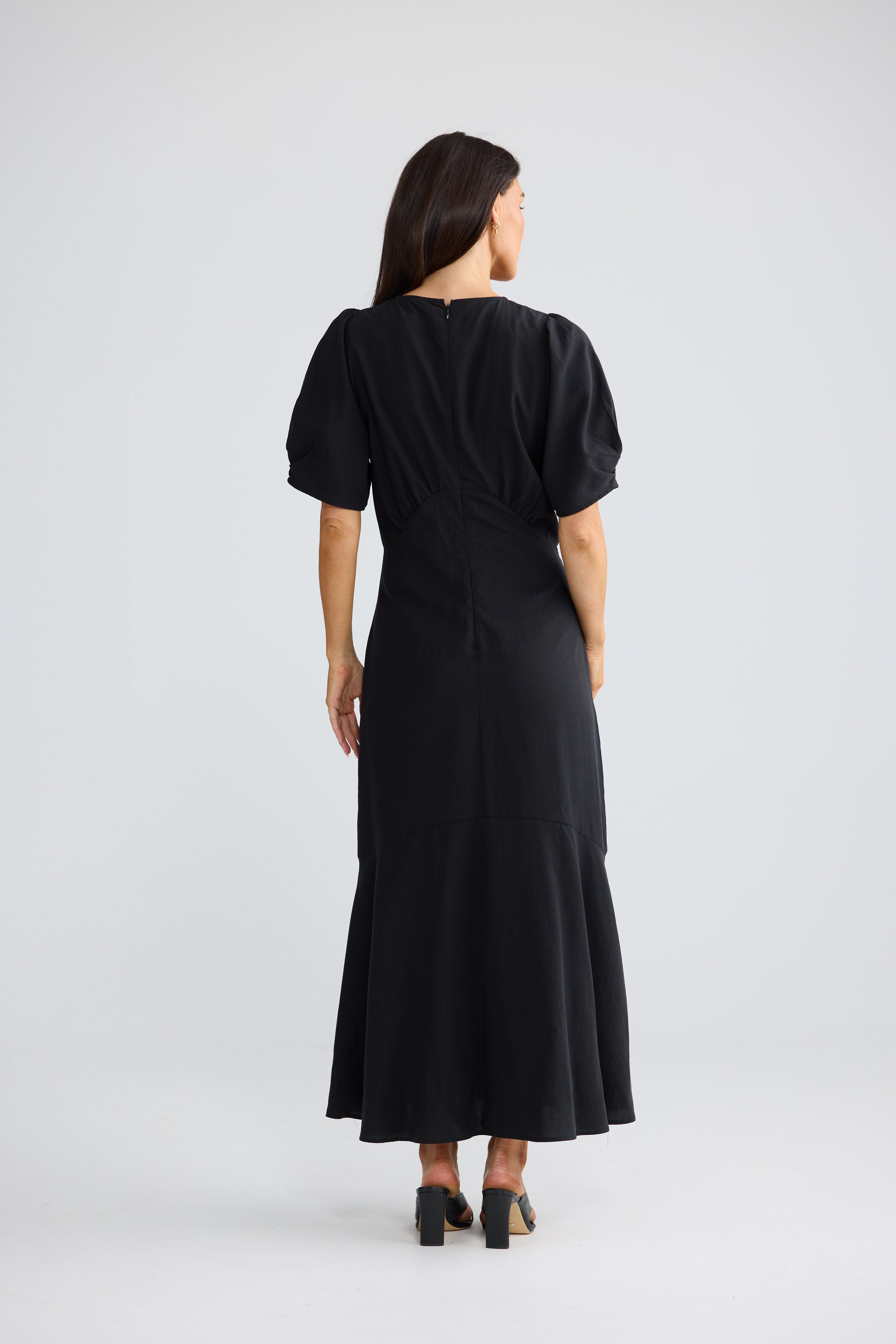 Image of A woman viewed from the back wearing a Brave & True black midi dress with short puff sleeves and a wrap-style back, standing against a light gray studio background.