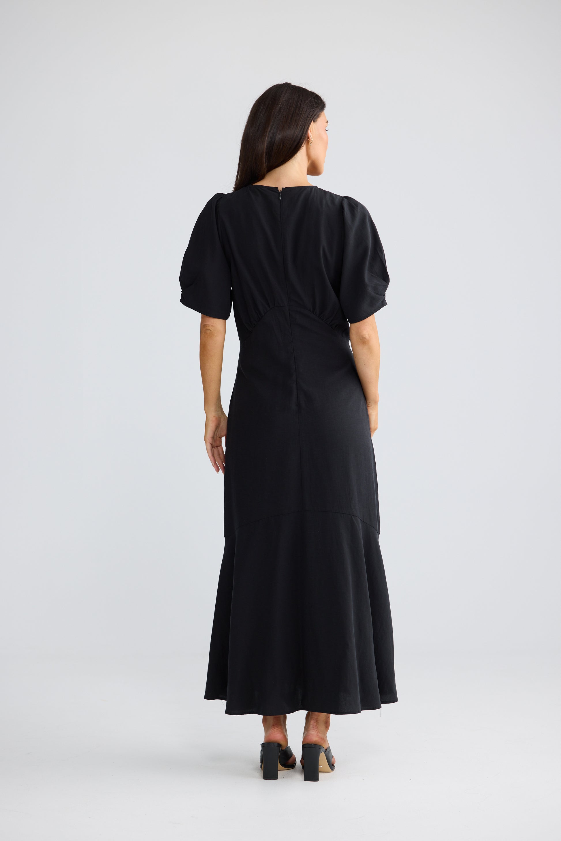 A woman viewed from the back wearing a Brave & True black midi dress with short puff sleeves and a wrap-style back, standing against a light gray studio background.