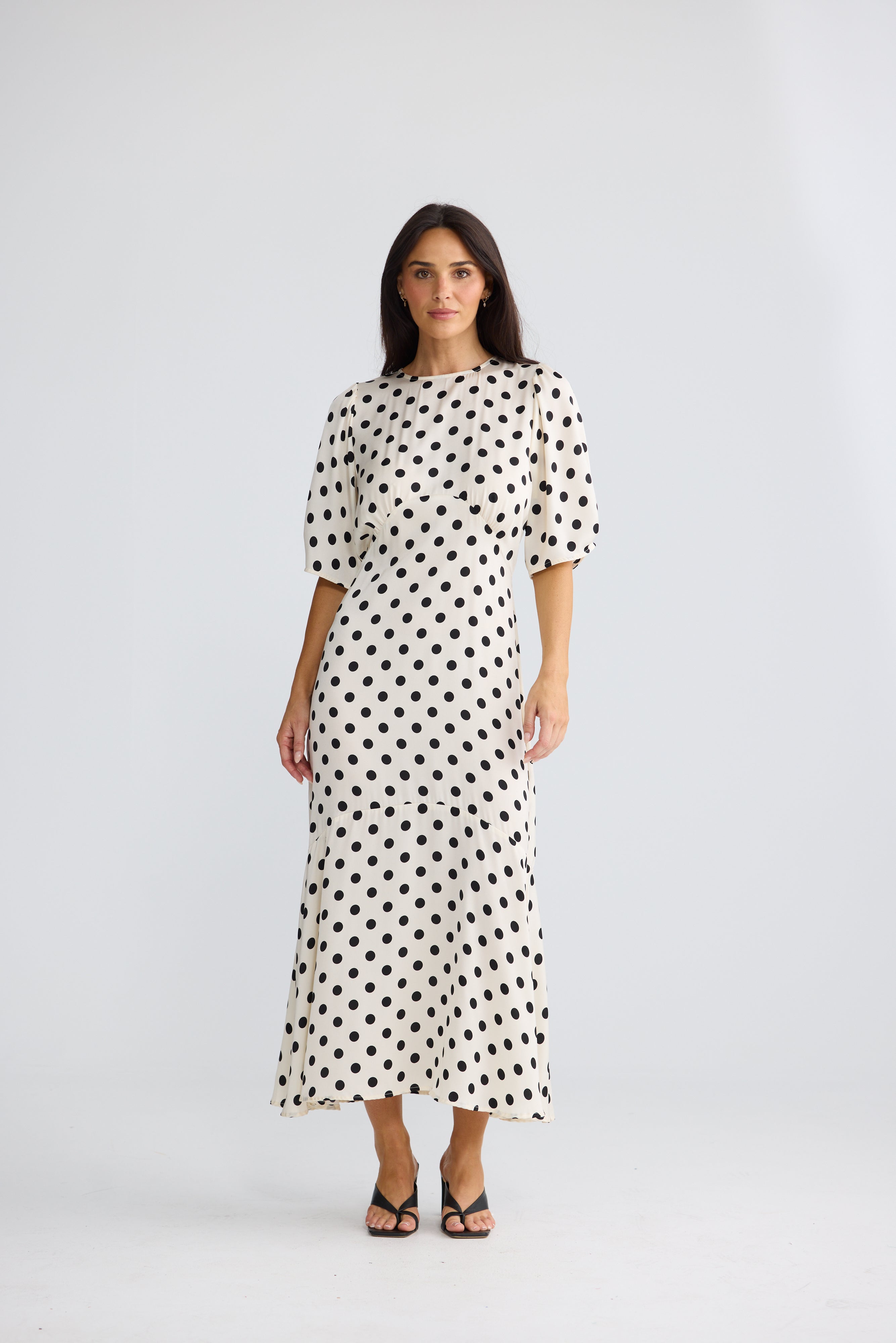 Image of A woman modeling a Brave & True midi-length cream dress with a black polka dot pattern, featuring short puff sleeves and a relaxed silhouette, set against a plain light gray studio background.