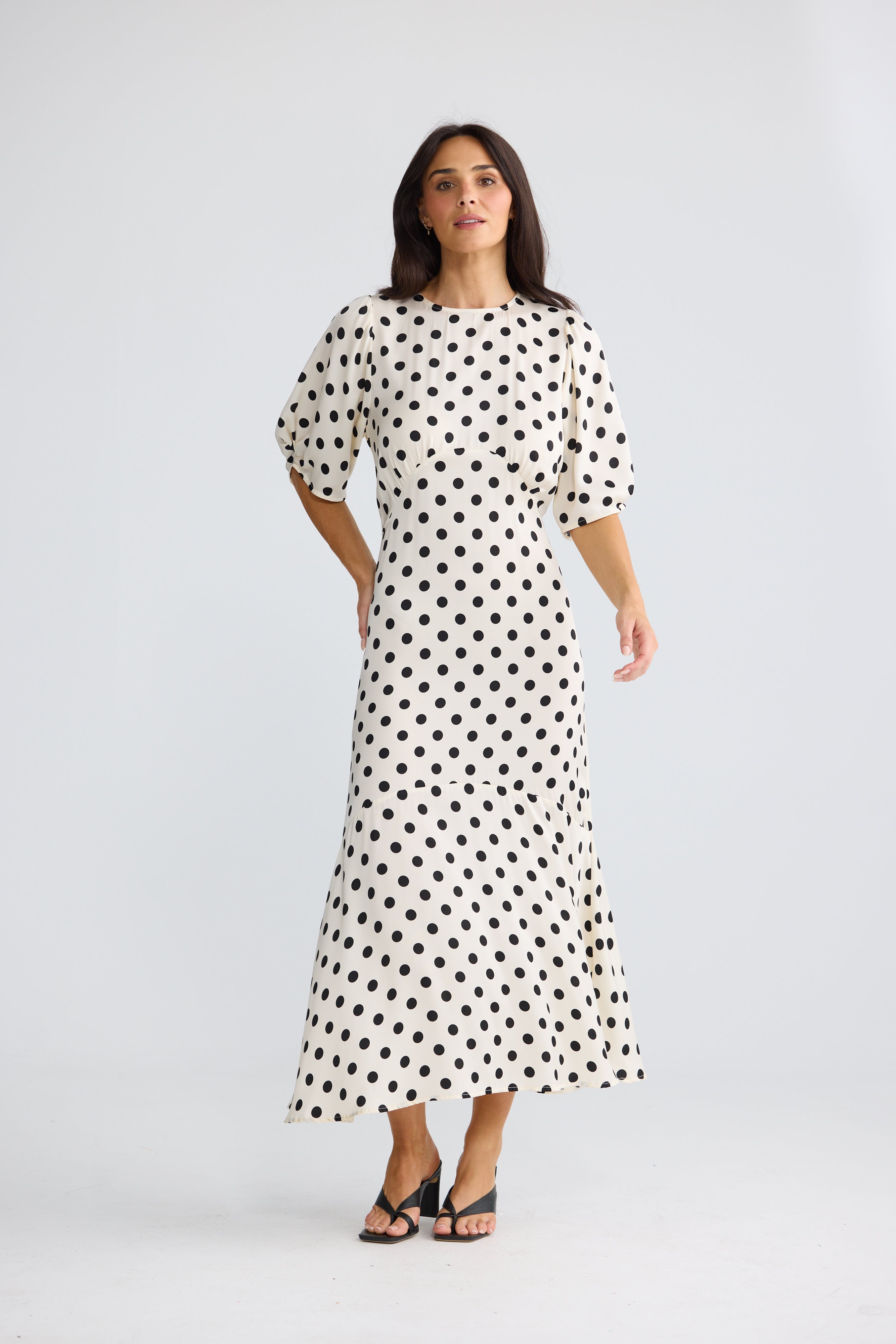 Image of A woman modeling a Brave & True white midi dress with black polka dots, featuring short puff sleeves and a boat neckline, set against a plain light gray studio background.