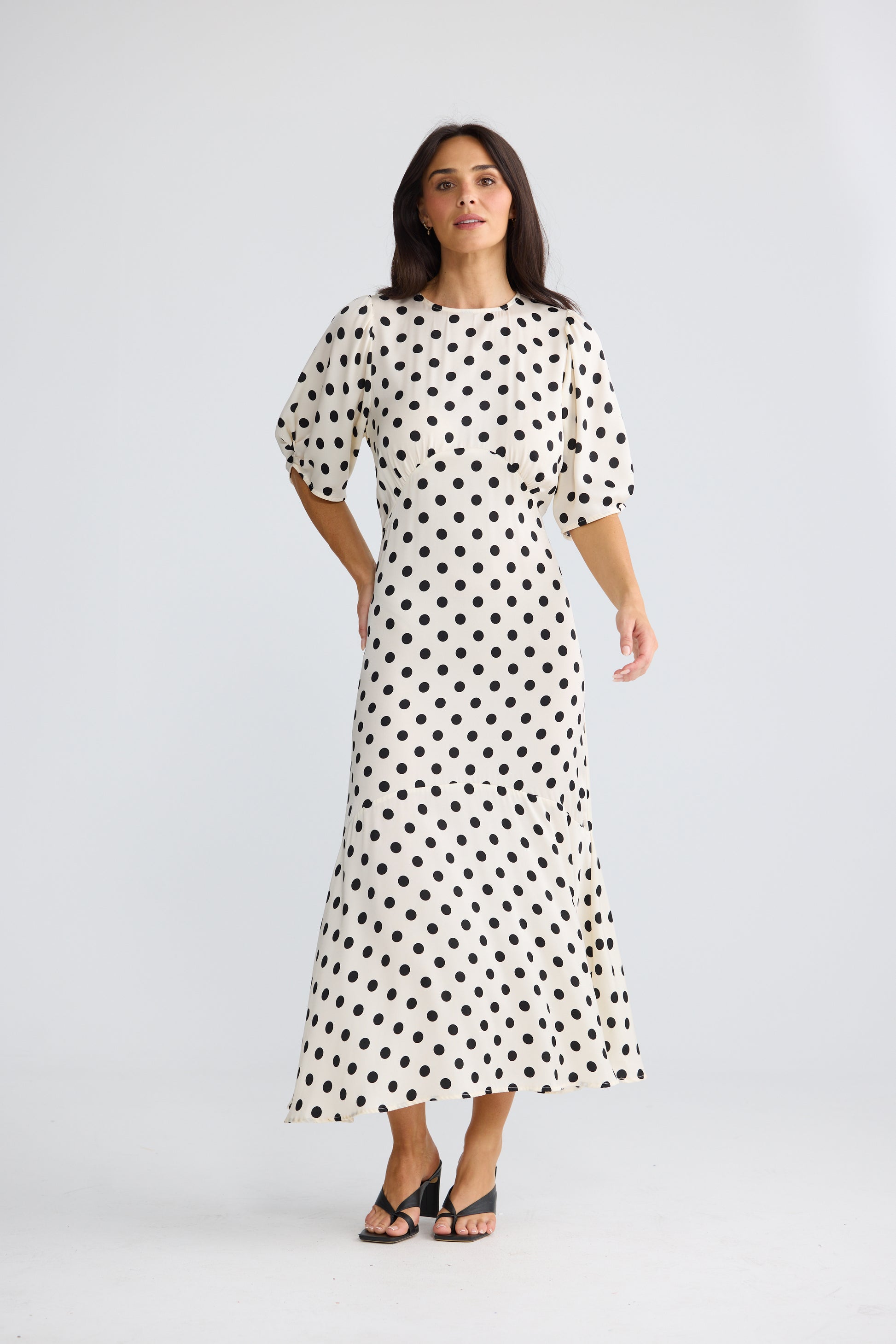A woman modeling a Brave & True white midi dress with black polka dots, featuring short puff sleeves and a boat neckline, set against a plain light gray studio background.