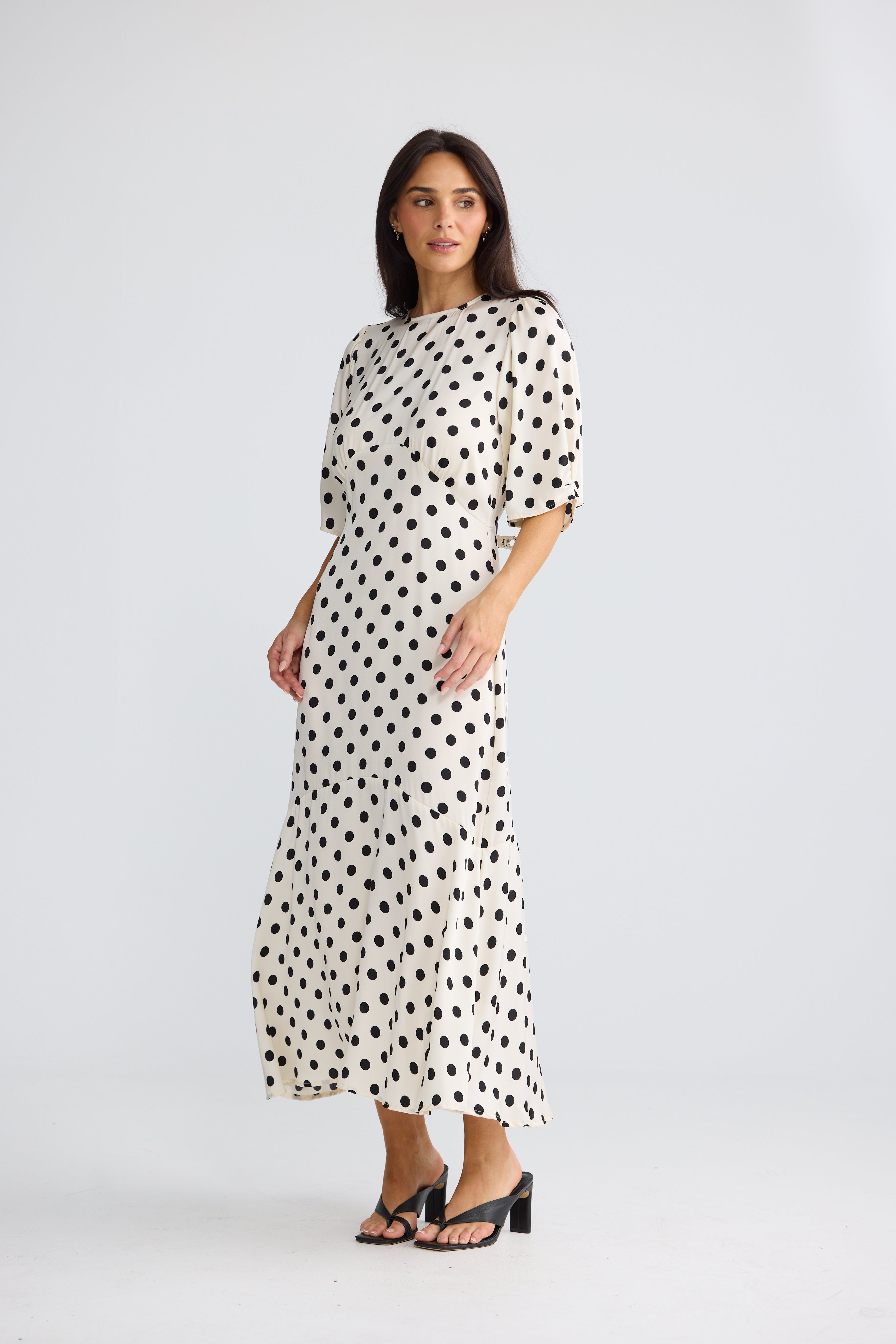 Image of A woman modeling a Brave & True midi-length cream dress with a black polka dot pattern, featuring elbow-length puff sleeves and a high round neckline, paired with black strappy heels against a light gray studio background.